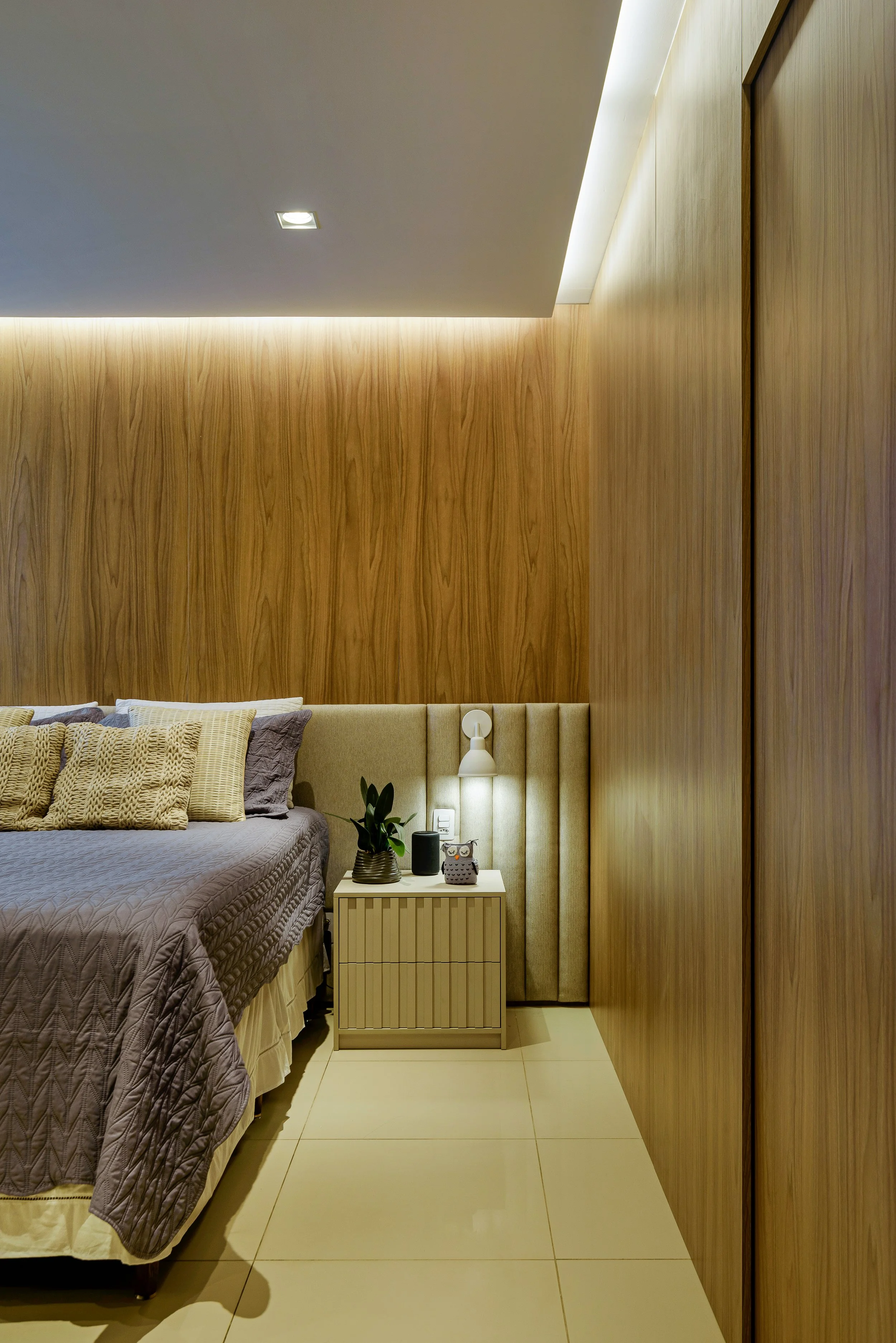 Interior of a bedroom with a wooden accent wall, bedside table with a plant, lamps, and decorative items, and a bed with pillows and a quilt.