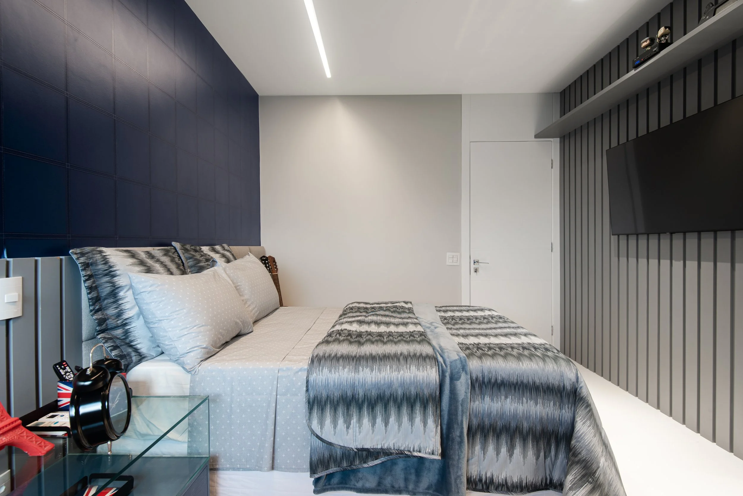 Modern bedroom with a bed, blue and gray bedding, gray paneling on the wall, mounted TV, and a glass bedside table.