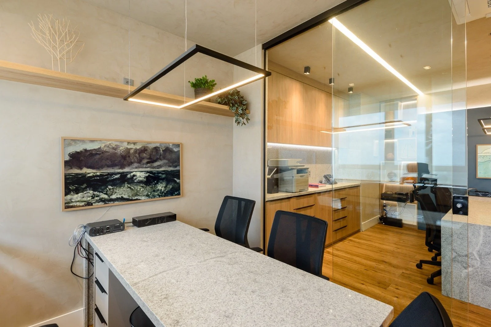 Office with a granite countertop table, black chairs, a framed landscape painting of a stormy sea, a small audio speaker, and a glass partition separating the room from a workspace with wooden cabinets, a desk, office chairs, and equipment.