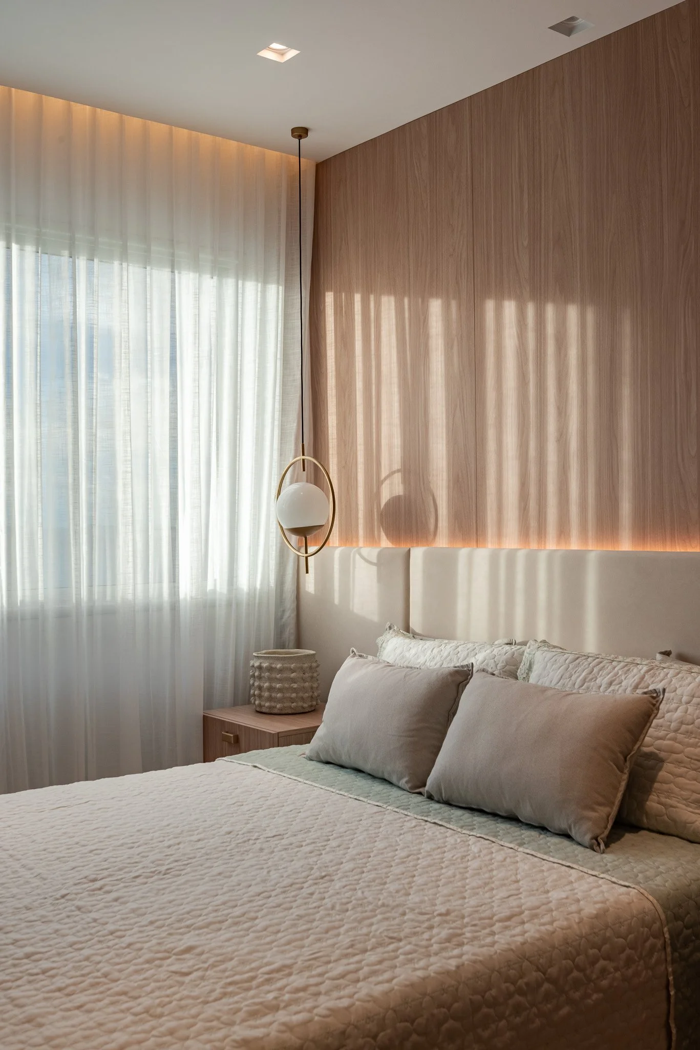 A modern bedroom with a bed, beige pillows, and a wooden wall behind the headboard. A bedside table with a textured ceramic lamp and a hanging pendant light near the window with sheer white curtains. The room has warm, natural lighting.