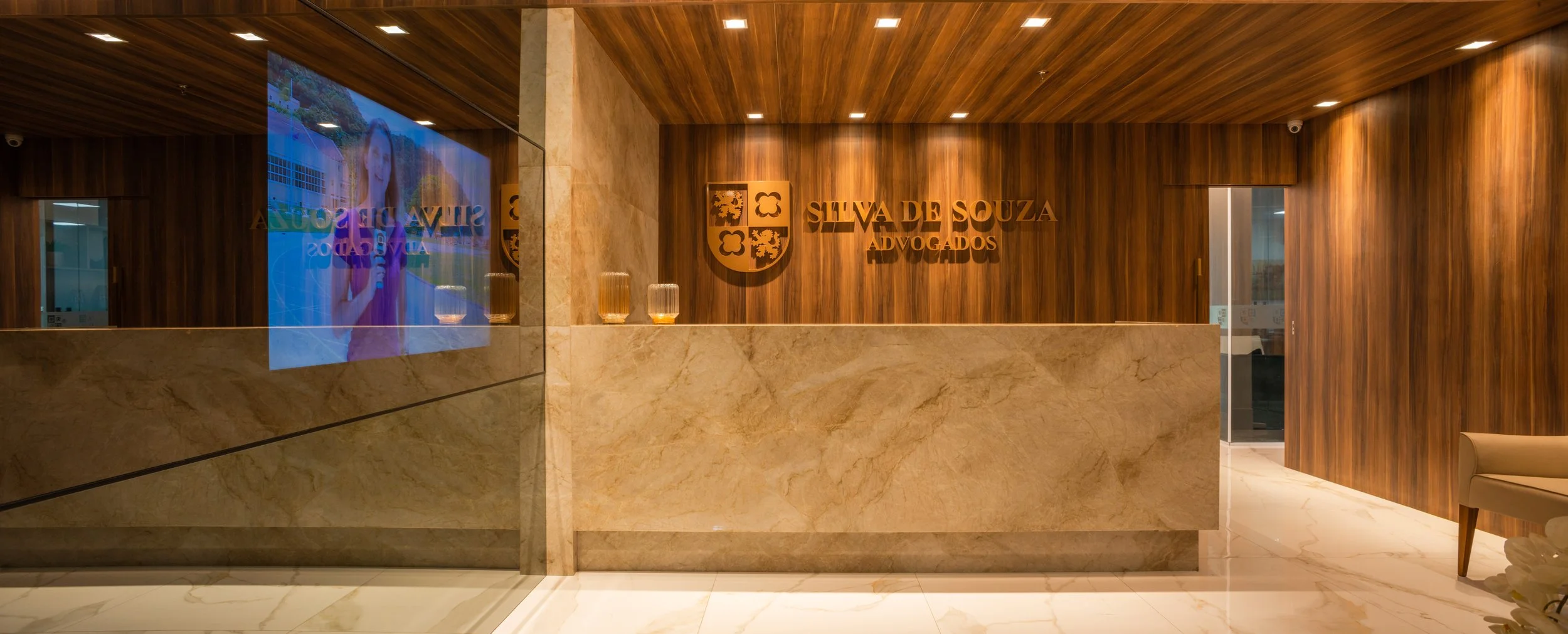 Reception area with wood-paneled walls, marble counter, logo of Silva de Souza Advogados, and a large mirror showing a woman speaking on TV.