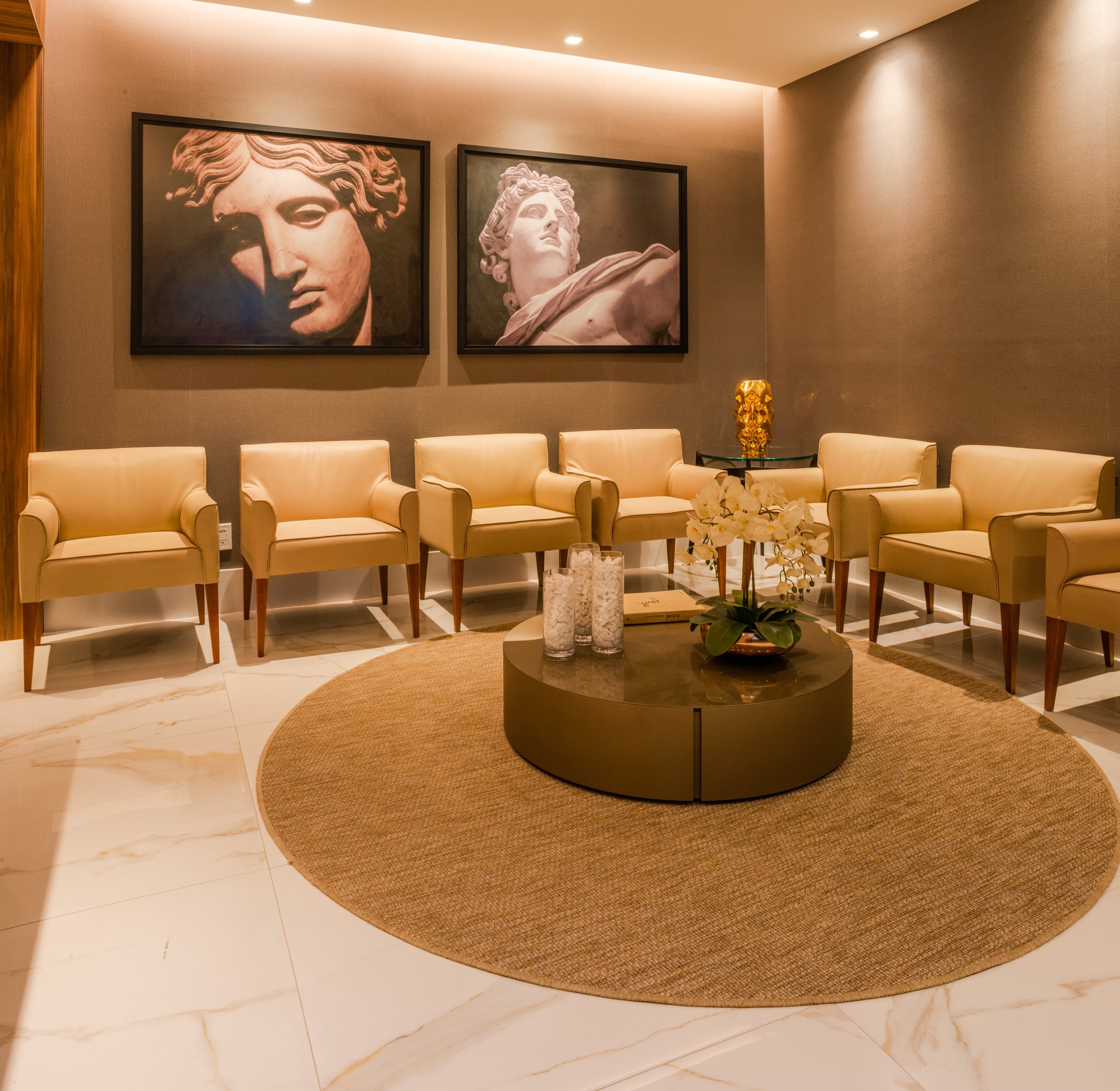 A waiting room with six beige armchairs arranged in a semi-circle around a round coffee table with a potted orchid plant and three glass vases. On the wall behind the chairs are two black-and-white framed photographs of classical sculptures, and a sm
