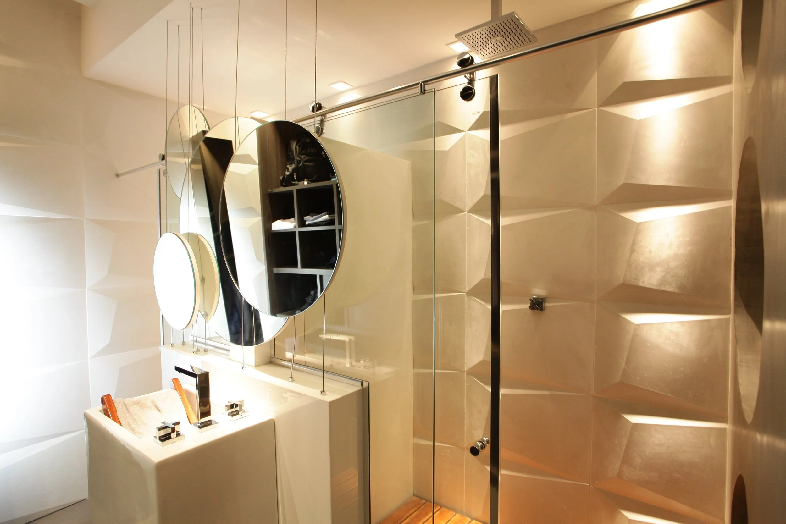 Modern bathroom with geometric wall tiles, large round mirrors, a glass shower door, and a white sink with a beige towel.