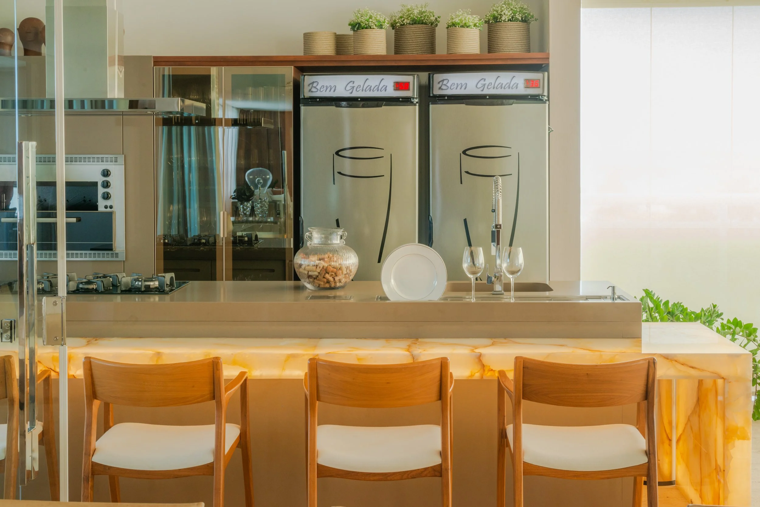 Modern kitchen with beige countertops, wooden chairs, two refrigerators, a glass jar with corks, a white plate, wine glasses, and green plants on top of the cabinets.