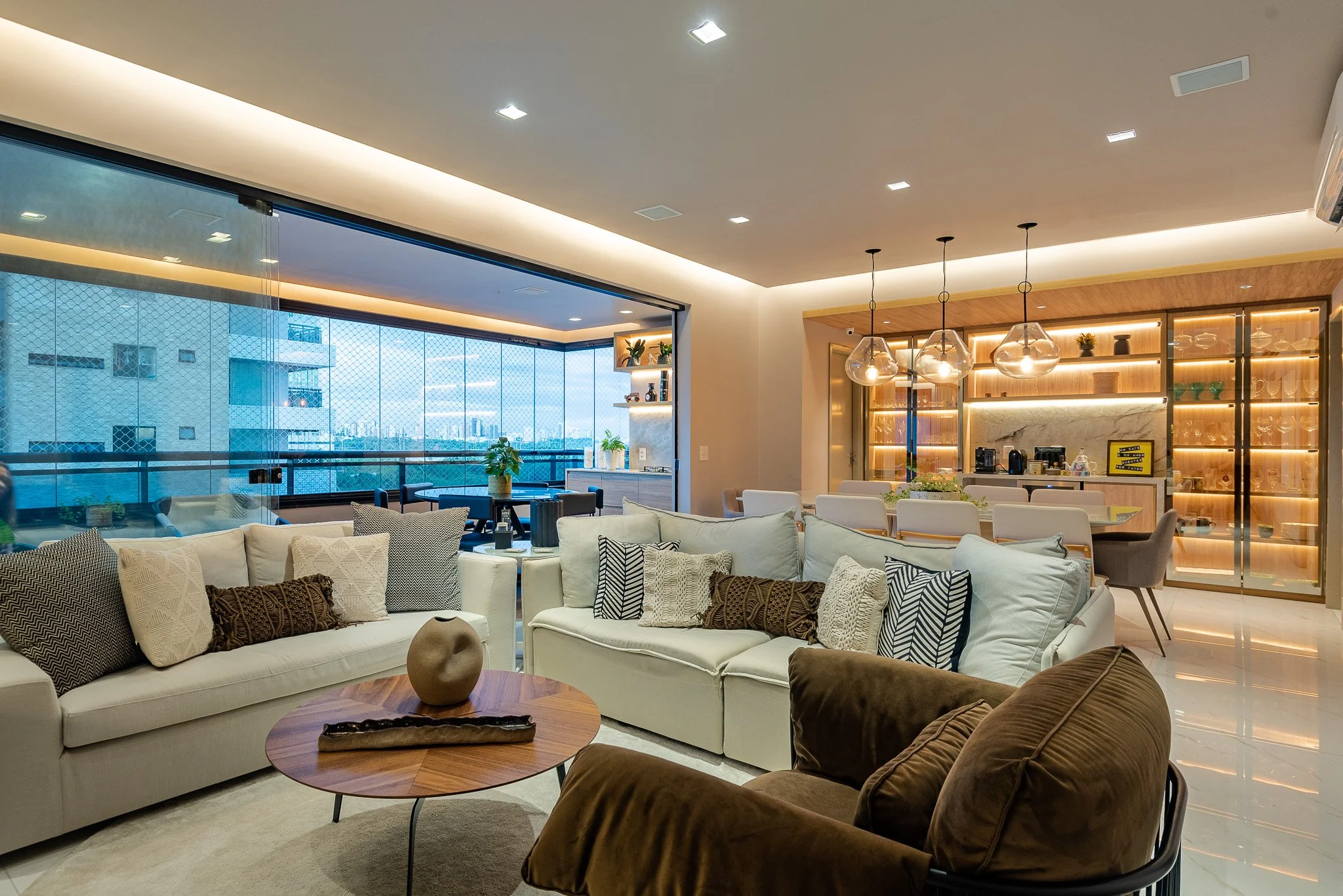 Modern living room with white sofas, brown armchair, round wooden coffee table, glass shelving with lighting, and city view balcony.