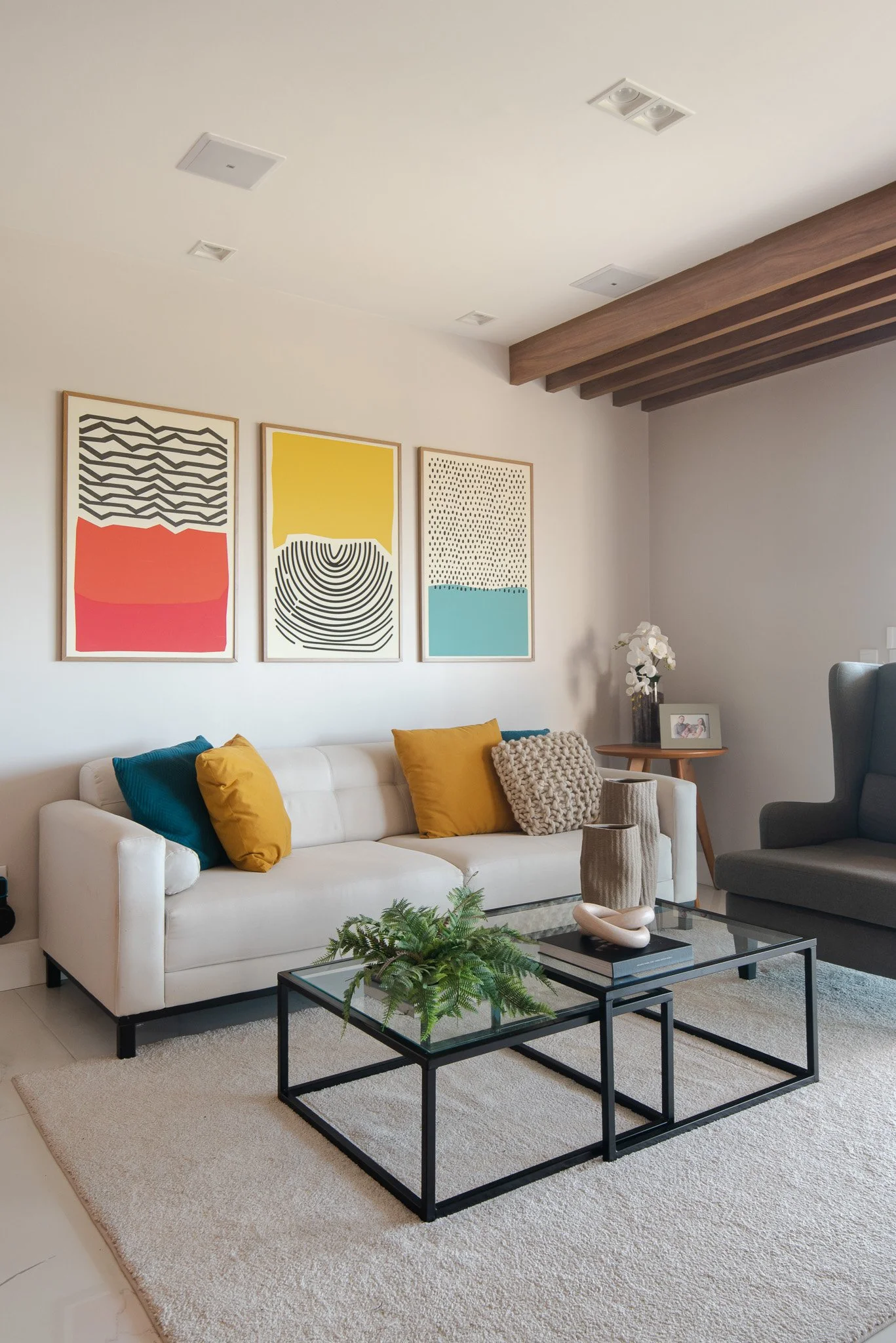 Living room with white sofa, yellow and blue pillow, abstract artwork on white walls, glass coffee table with decorative objects, potted plant, and side table with flowers and framed photo.