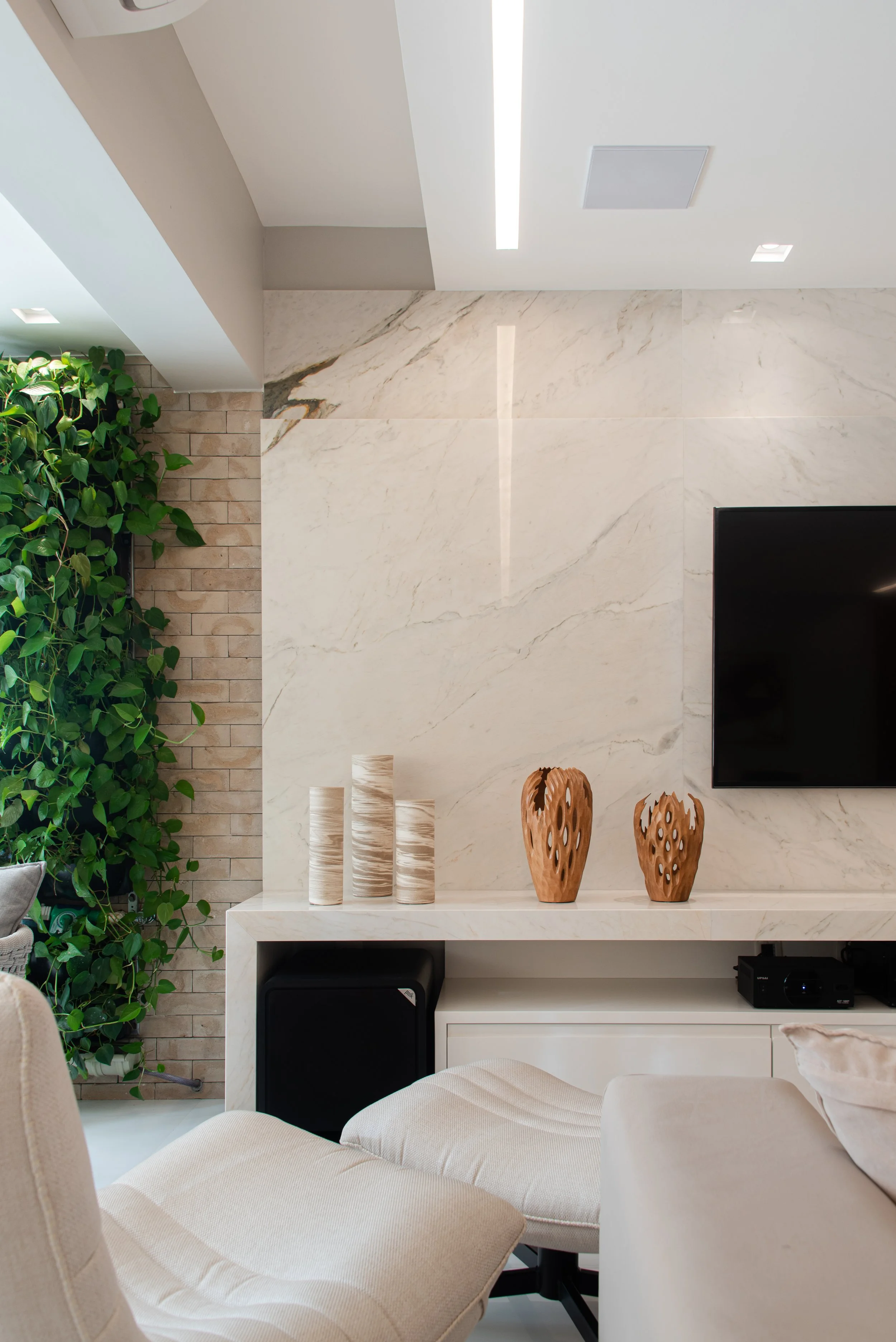 Living room with beige cushioned chair, decorative vases on white shelf, marble wall, TV, green plant on brick wall, ceiling lighting.