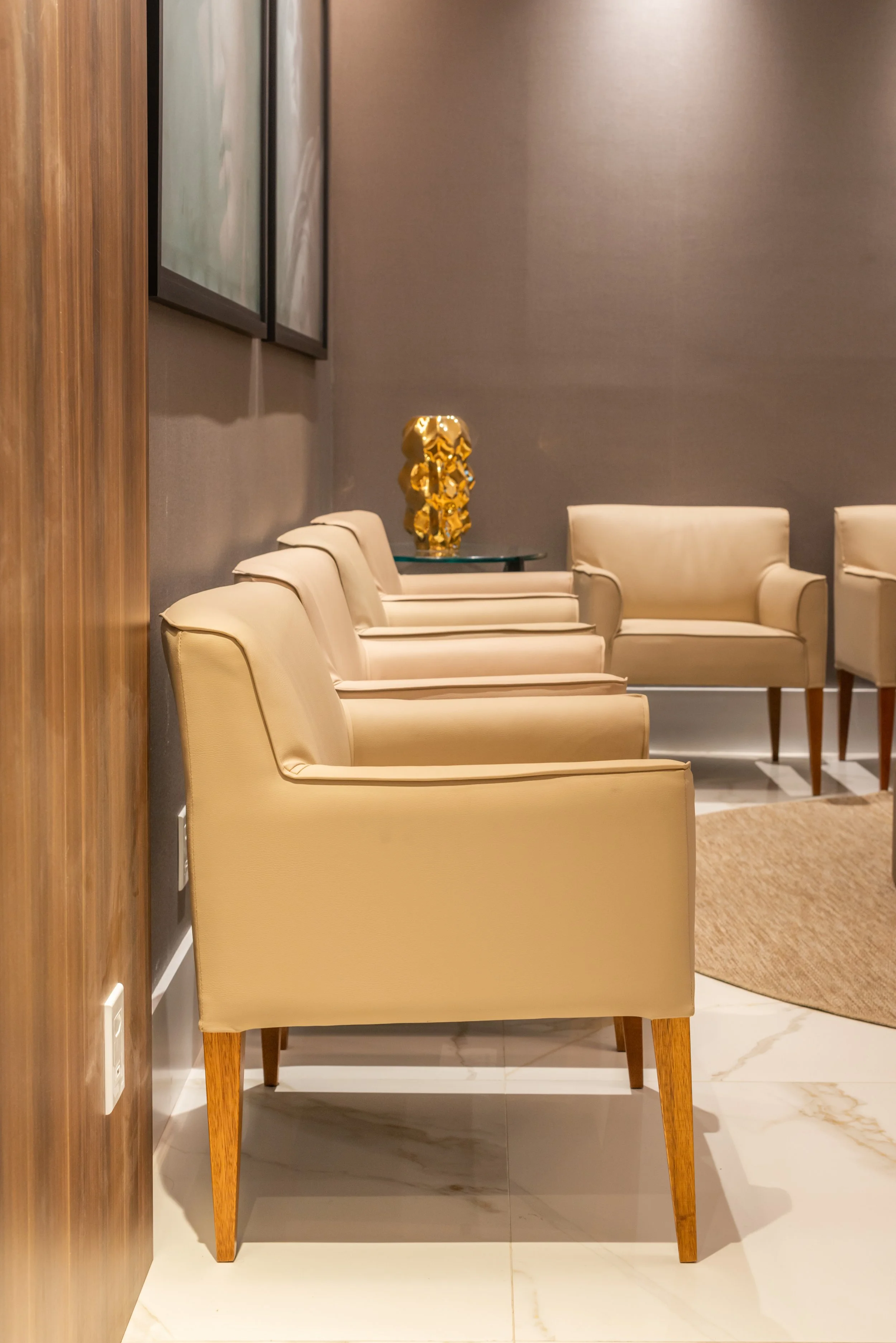 A row of cream-colored armchairs with wooden legs in a modern hotel lobby, with a gold decorative sculpture on a side table and framed artwork on the wall.
