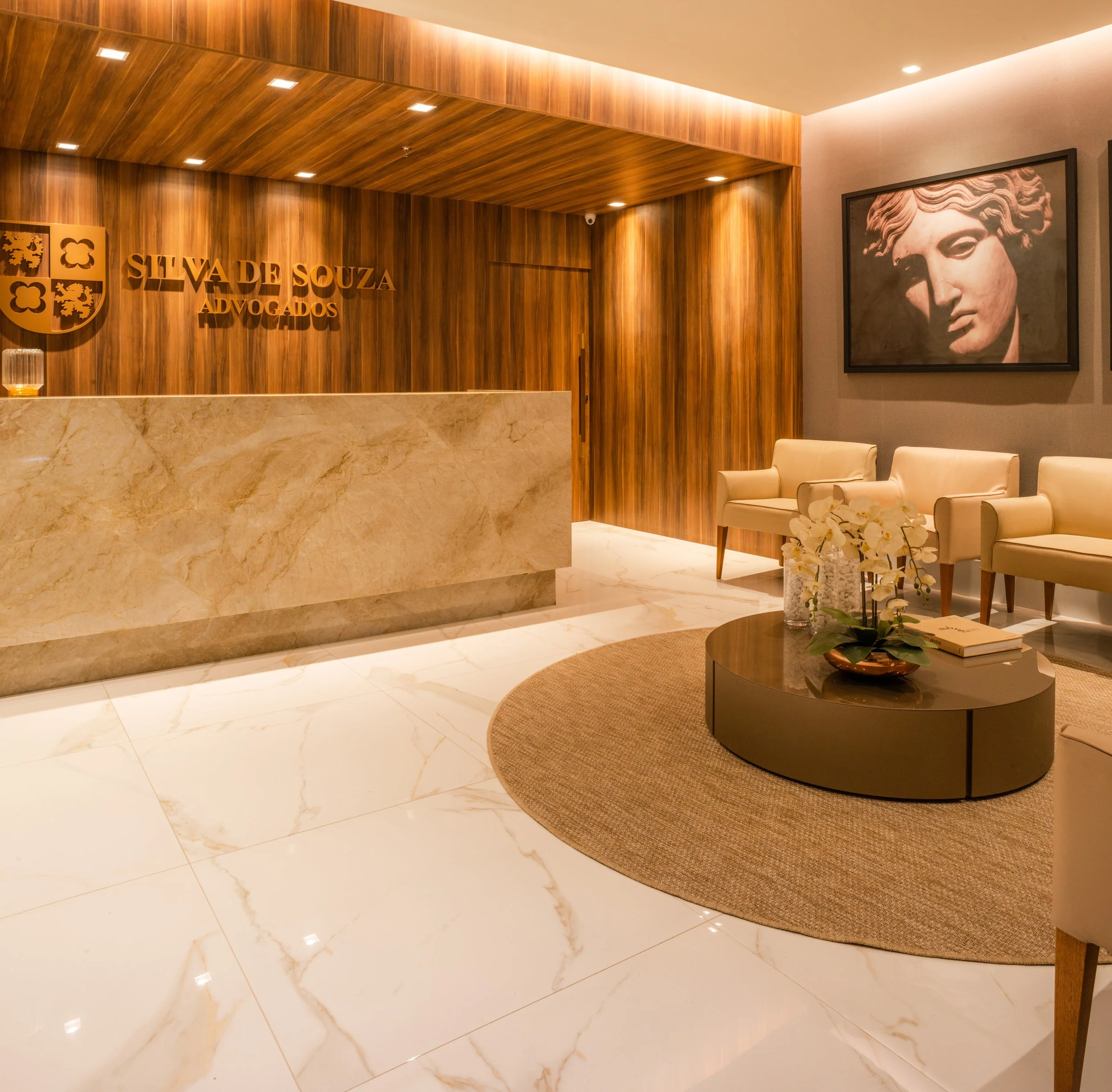 Reception area with wooden wall paneling, a beige marble counter, and a sign that reads 'Silva de Souza Advogados.' Modern waiting area with cream-colored chairs, a coffee table with white orchids, a book, and a framed black-and-white portrait of a c