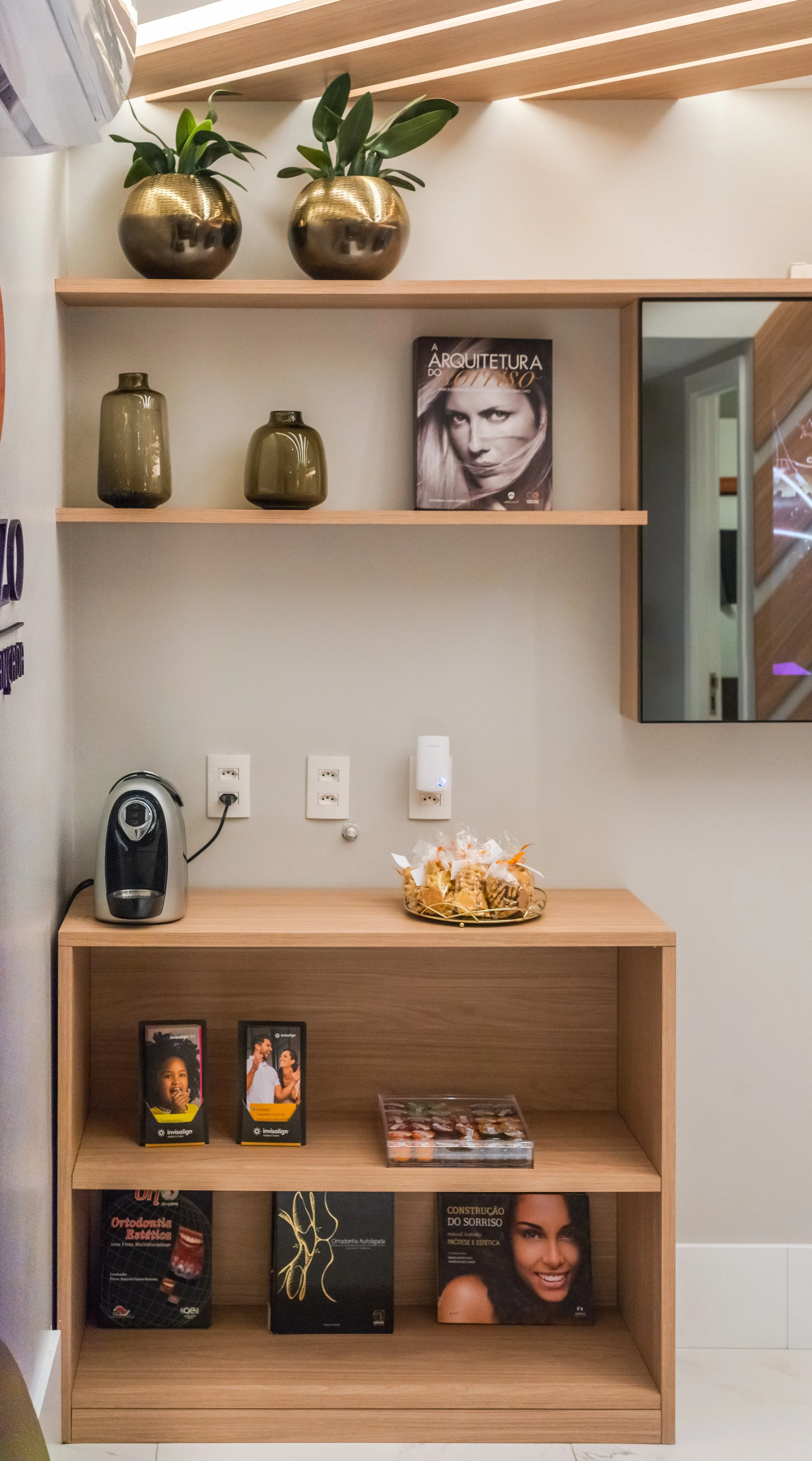 Wooden shelf unit with decorative plants, vases, a magazine, a mirror, a coffee machine, snacks, and reading materials on the shelves.