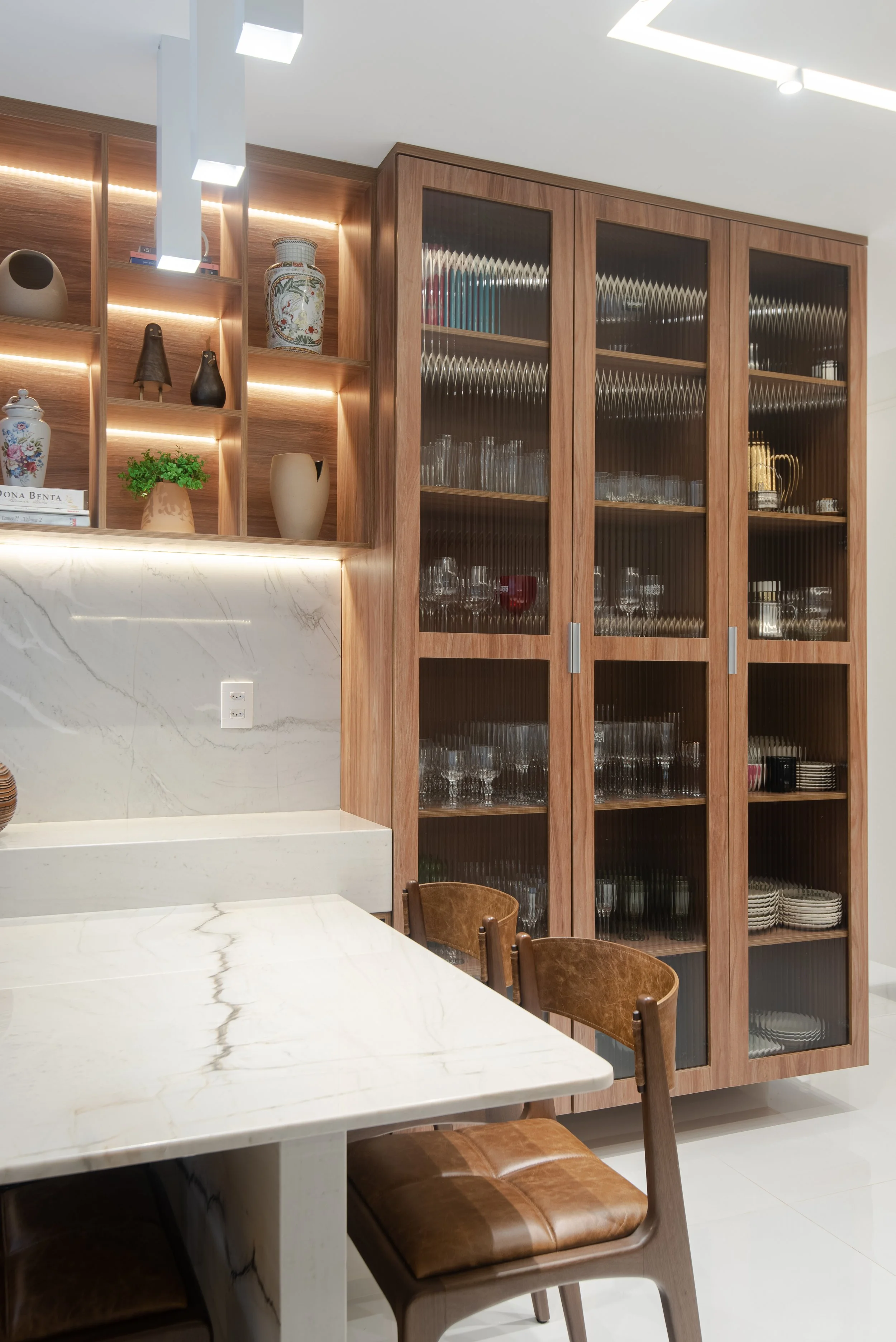 A modern interior with a marble table, wooden chairs, a large wooden cabinet with glass doors filled with glassware, and open shelves with decorative items and plants.
