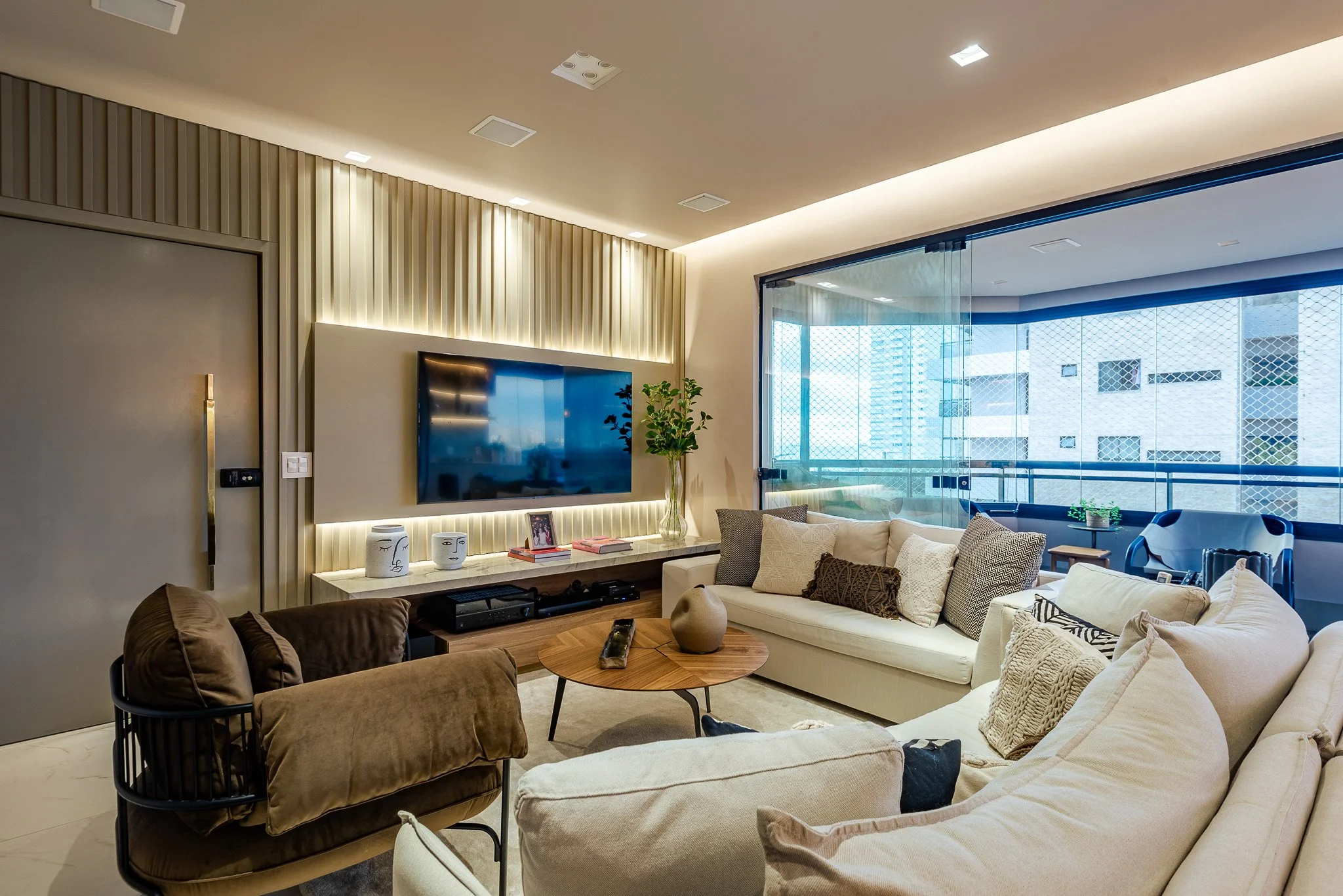 Modern living room with beige sectional sofa, brown armchair, wooden coffee table, and wall-mounted TV, large glass sliding door with city view in background, decorative pillows and plants.