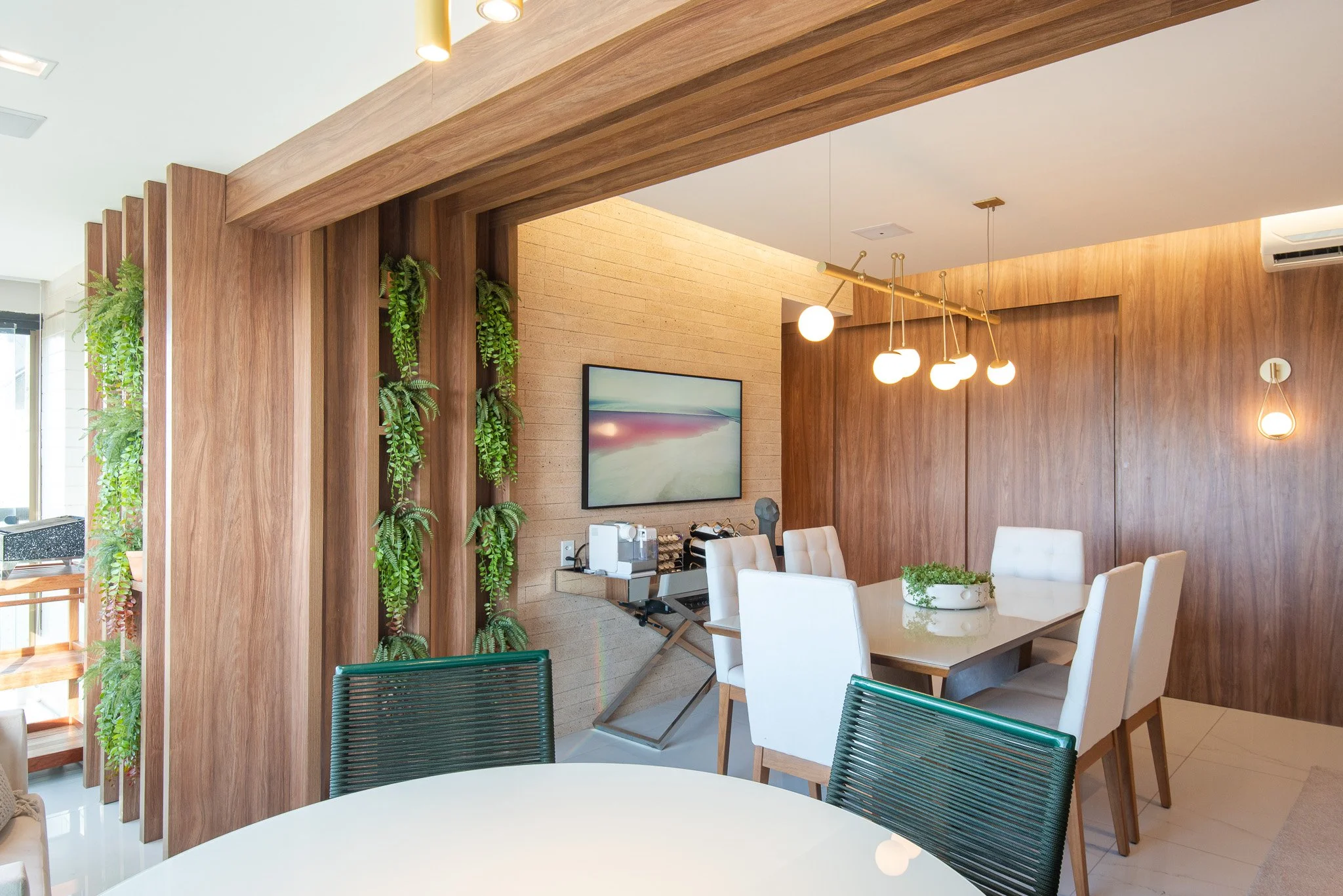 Modern dining room with wooden walls, white chairs around a rectangular table with a plant centerpiece, hanging globe lights, a wall-mounted TV, and indoor plants.