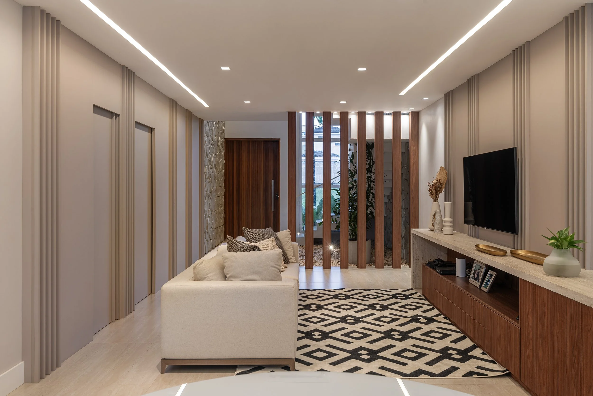Modern living room with a white sofa, patterned rug, wooden and stone wall accents, vertical wooden slats, and a mounted flat-screen TV.