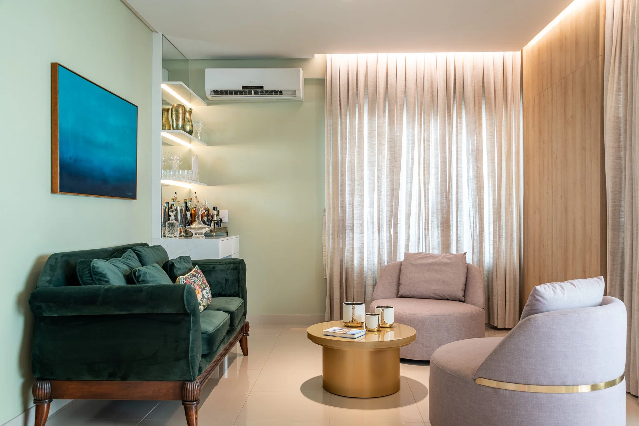 A modern living room with a dark green velvet sofa, a round gold coffee table with three white and gold cups, two light-colored armchairs, a large window with beige curtains, wooden paneling on the wall, a mounted air conditioning unit, and a white s