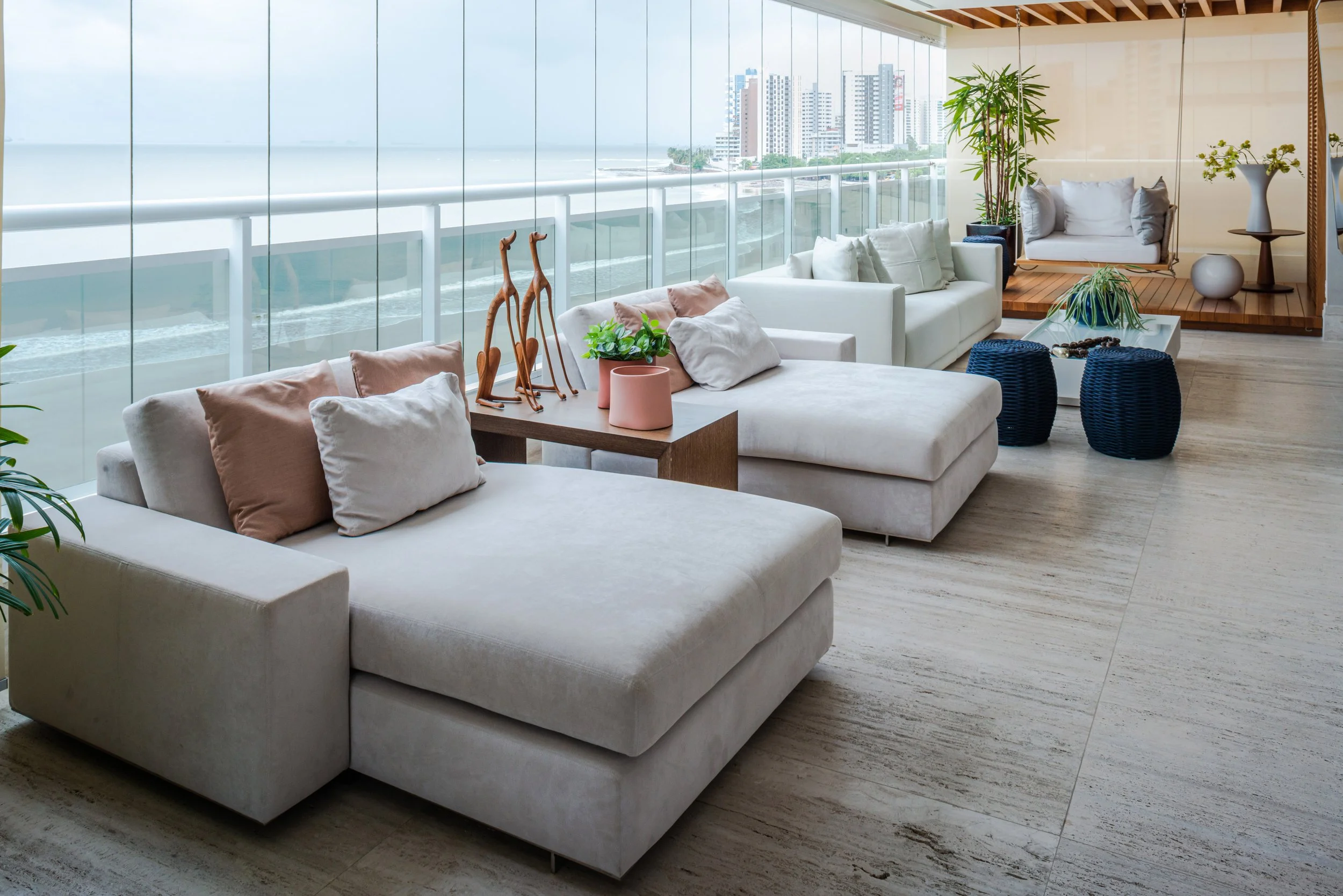 Luxurious living room with white sectional sofas, decorative pillows, plants, and a glass wall overlooking the ocean and city skyline.