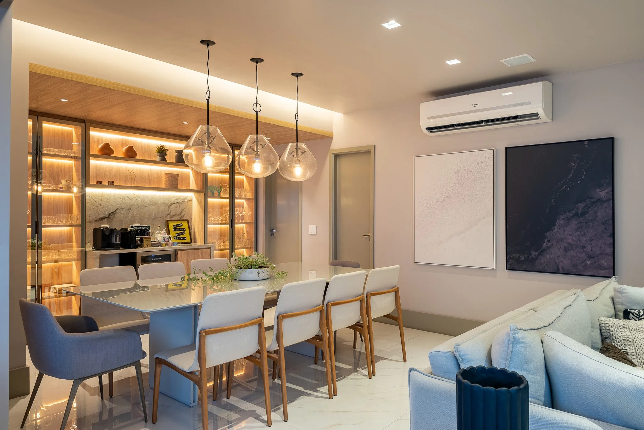Modern living room and dining area with a white sofa, abstract artworks on the wall, a dining table with eight chairs, pendant lights, and a built-in wooden and marble bar area.