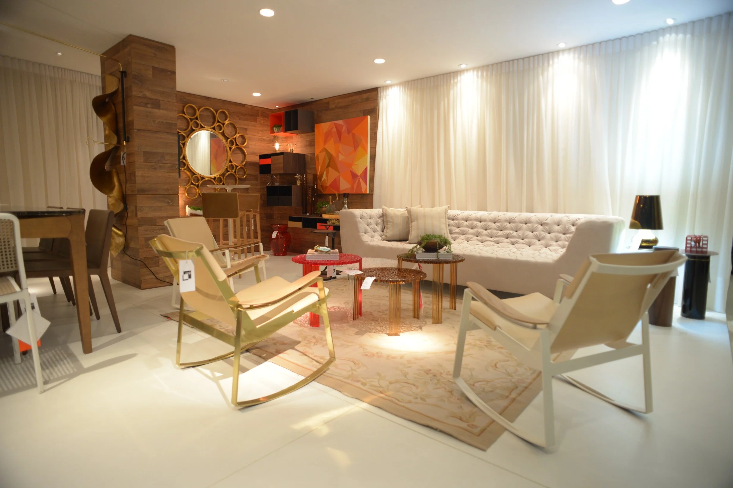 A living room with a white tufted sofa, a patterned rug, and several chairs including cream and gold rocking chairs, with round coffee tables. The room features a wooden accent wall with a mirror and colorful abstract art, decorated with vases and li