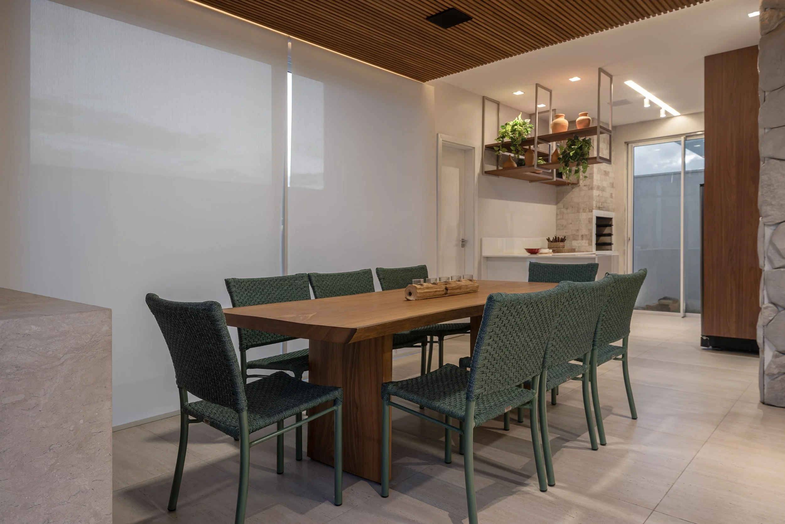 Modern dining room with a wooden table surrounded by six green upholstered chairs, a stone and wooden ceiling, a wall with floating shelves holding plants and pottery, a built-in outdoor fireplace, and glass sliding doors leading outside.