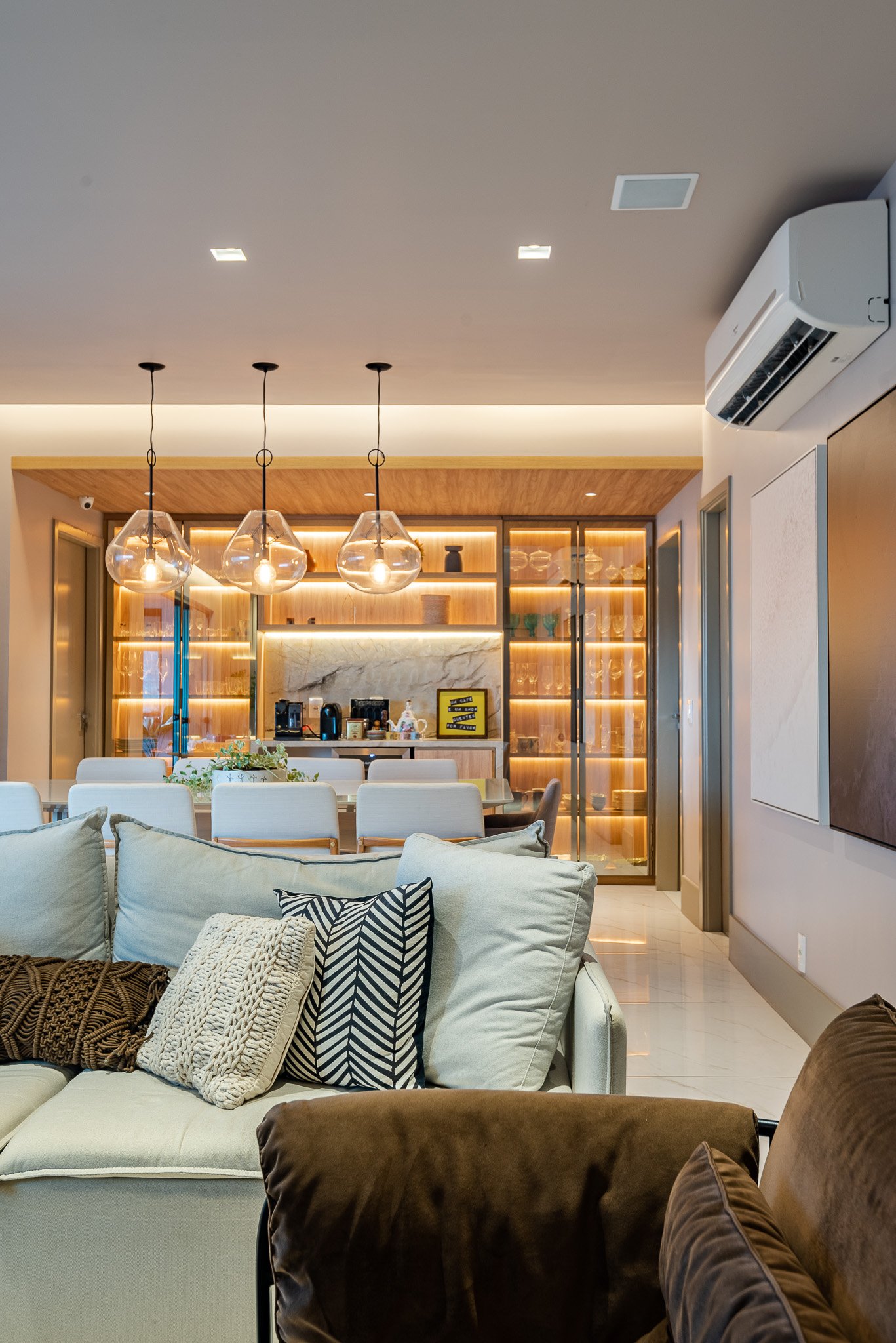 Modern living room and dining area with beige sofa, decorative pillows, a glass-fronted display cabinet, and pendant lighting.
