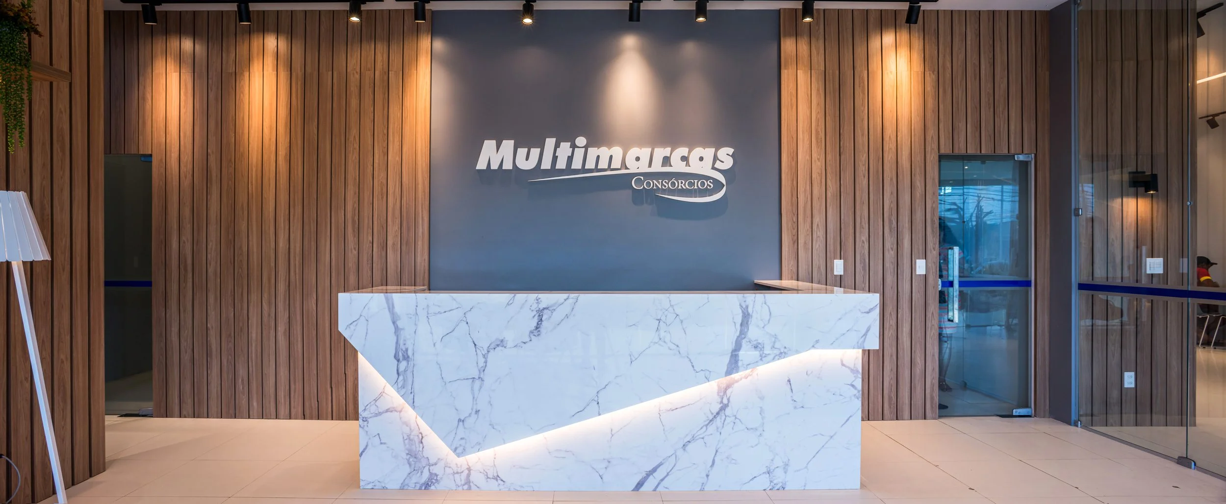 Modern office reception desk with marble top, wooden panel walls, and a blue logo reading 'Multimargas Consórcios' on the wall behind the desk.