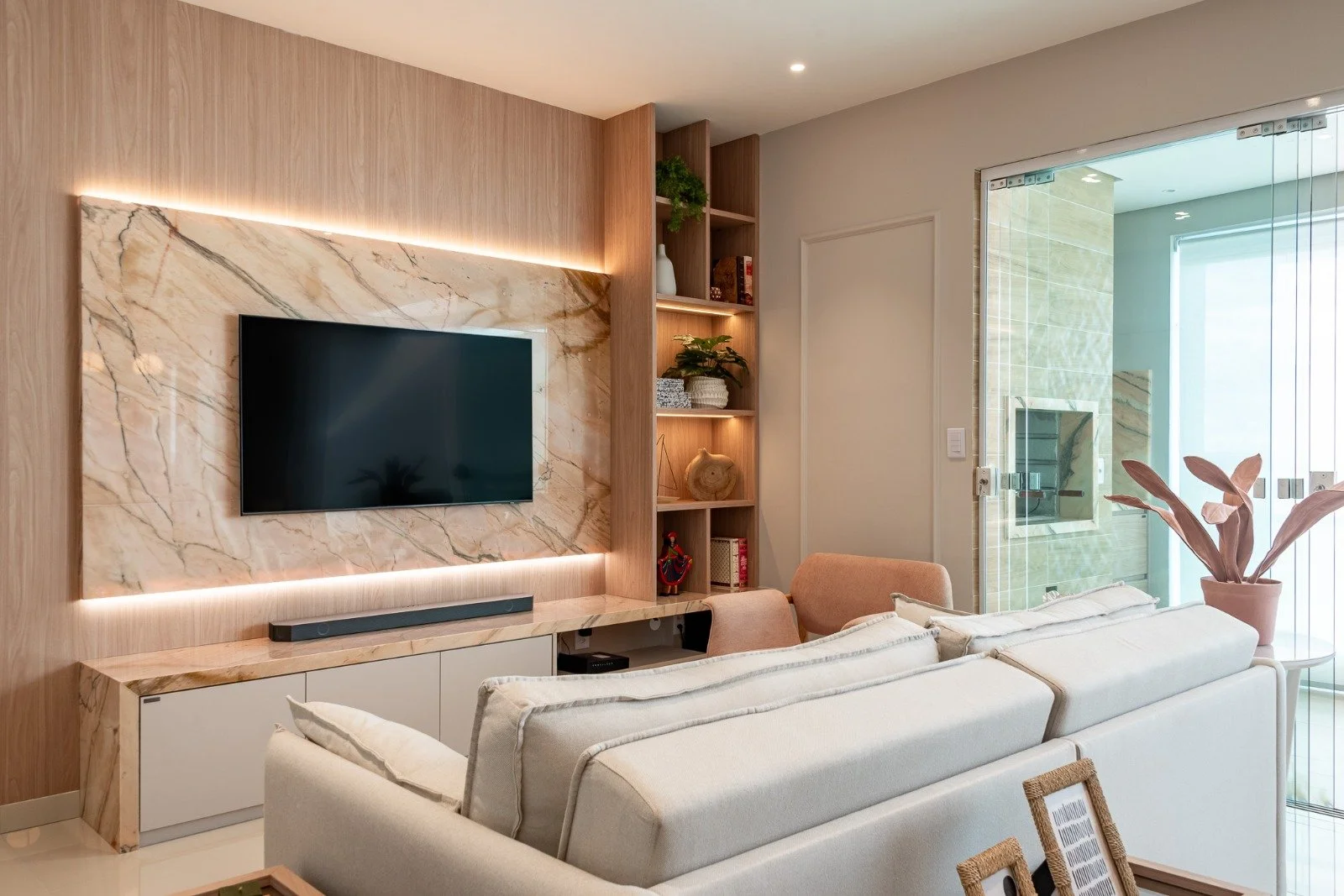 Living room with a wall-mounted TV on a marble accent wall, light wood paneling, and decorative shelving filled with plants and decor, adjacent to a glass door leading to a balcony with a fireplace.