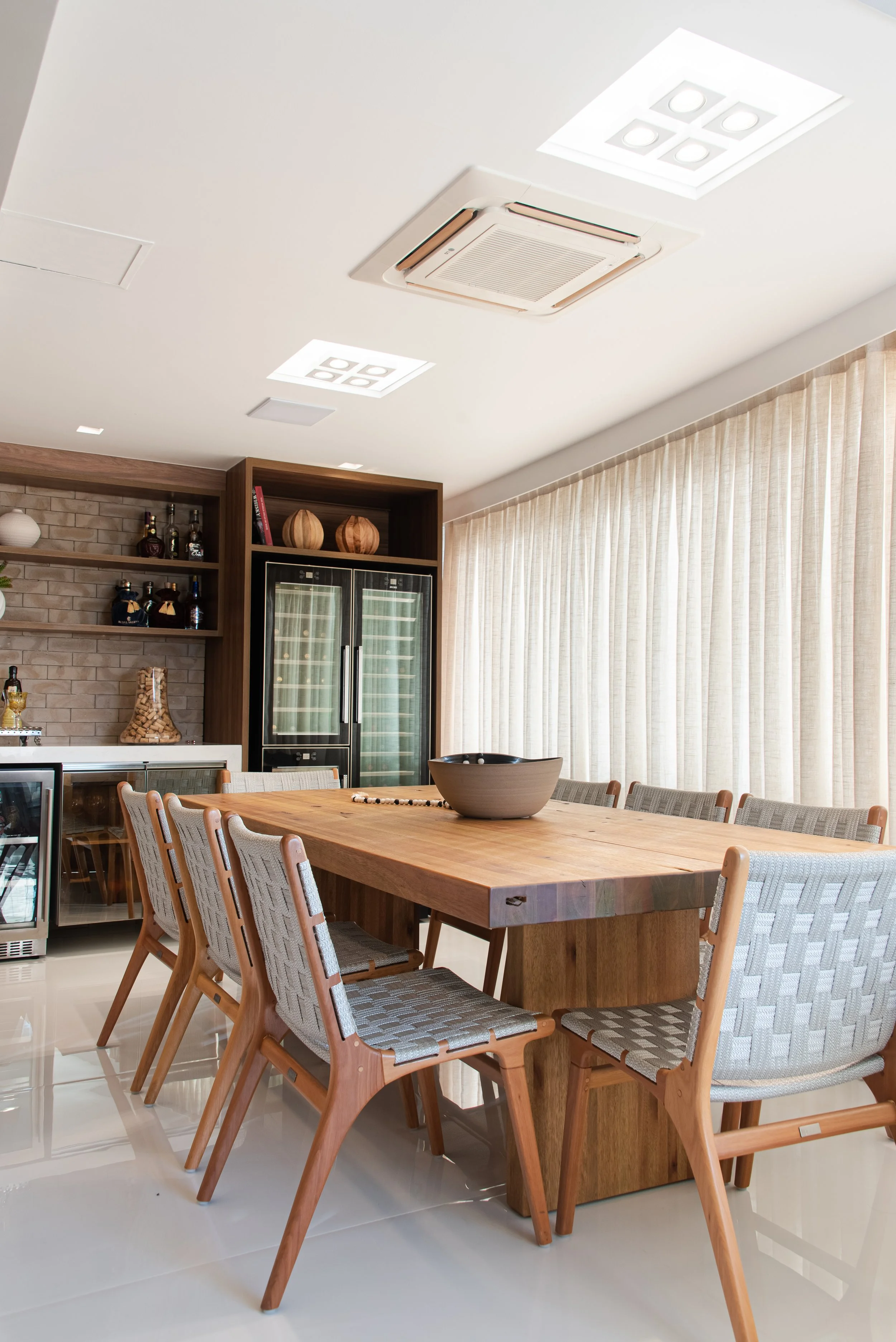 Modern dining room with a large wooden table surrounded by six wooden chairs with woven fabric seats, beige curtains covering large windows, a beige brick wall with shelves, and a wine cooler.