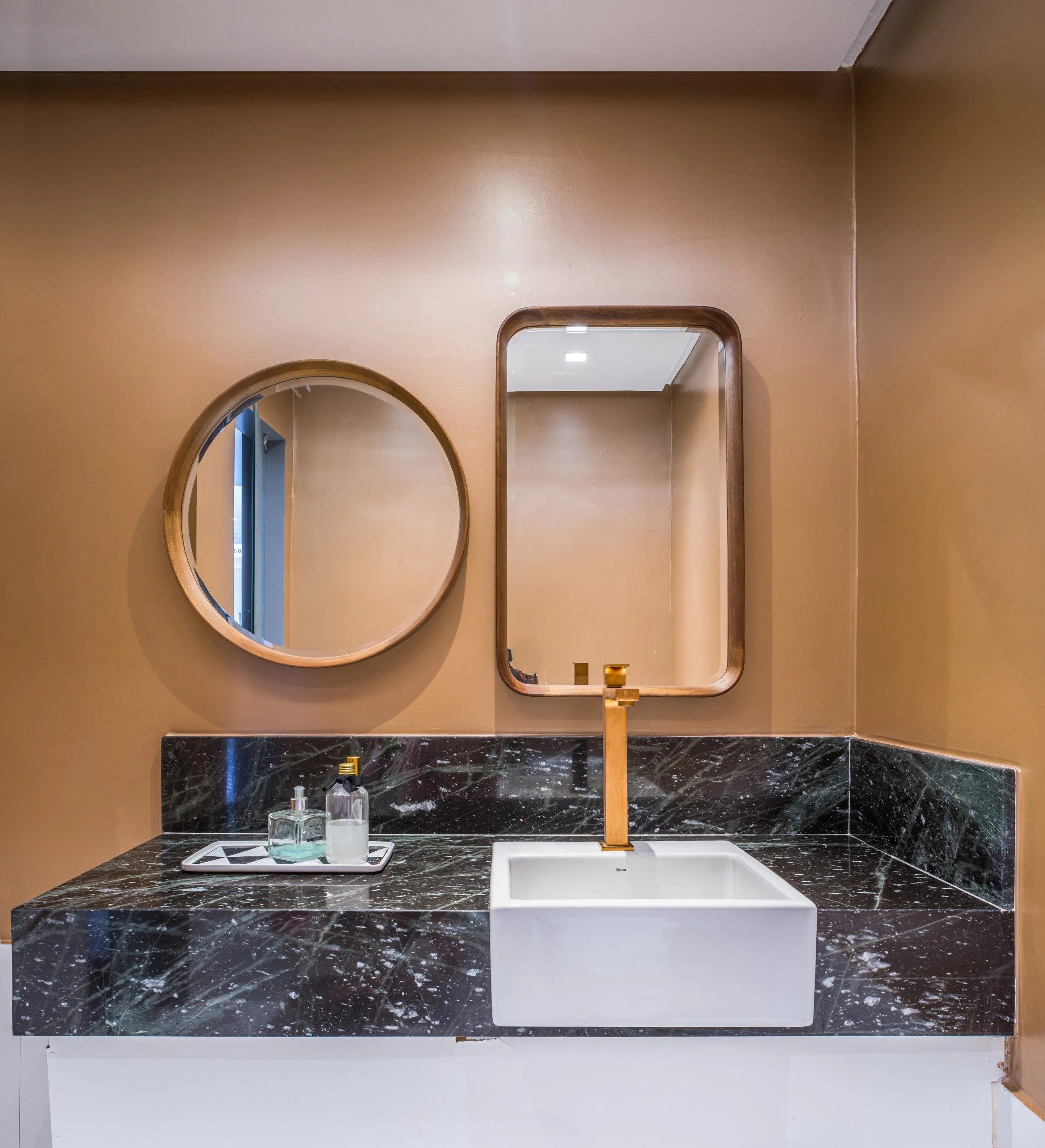 Modern bathroom vanity with a black marble countertop, a white rectangular sink, a gold faucet, two mirrors (one round and one rectangular), and a tray with toiletries on a brown wall background.