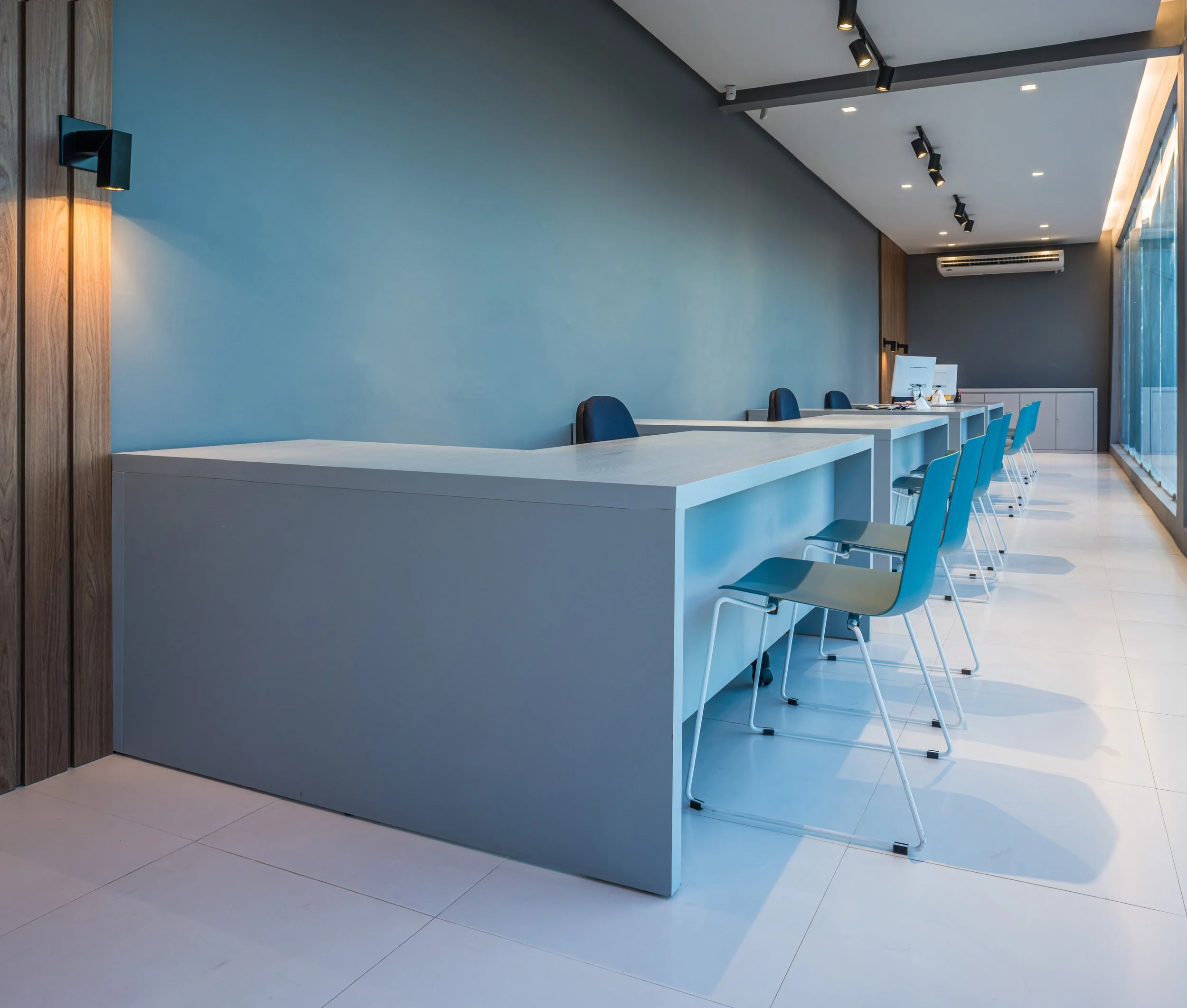 Empty modern office space with a white desk, teal chairs, and computer stations near large windows.