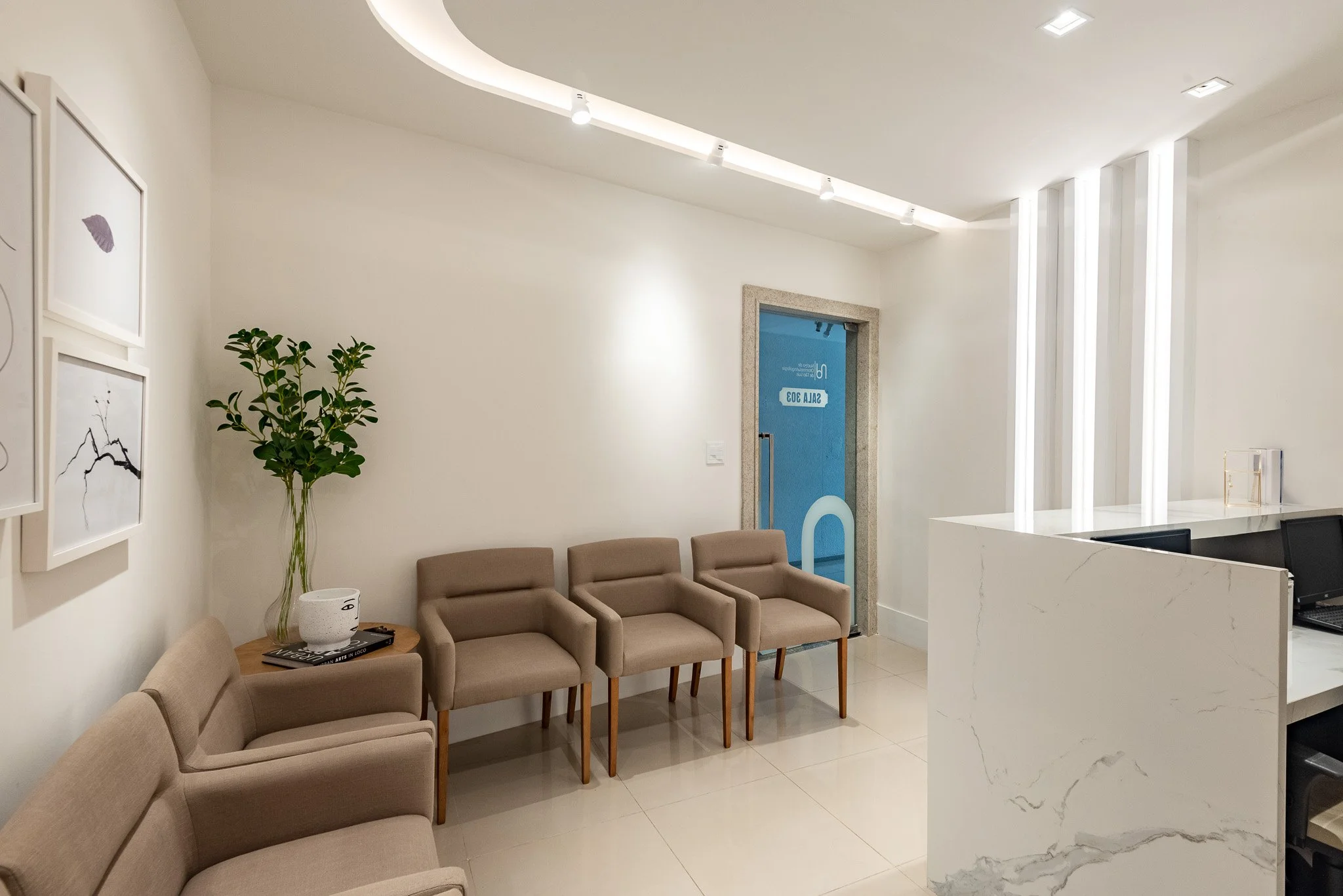 Modern waiting room with beige chairs, a large potted plant, and minimalist artwork on white walls. A reception desk with a white marble top and black chairs is visible. The room is well-lit with ceiling lighting and vertical LED light fixtures.