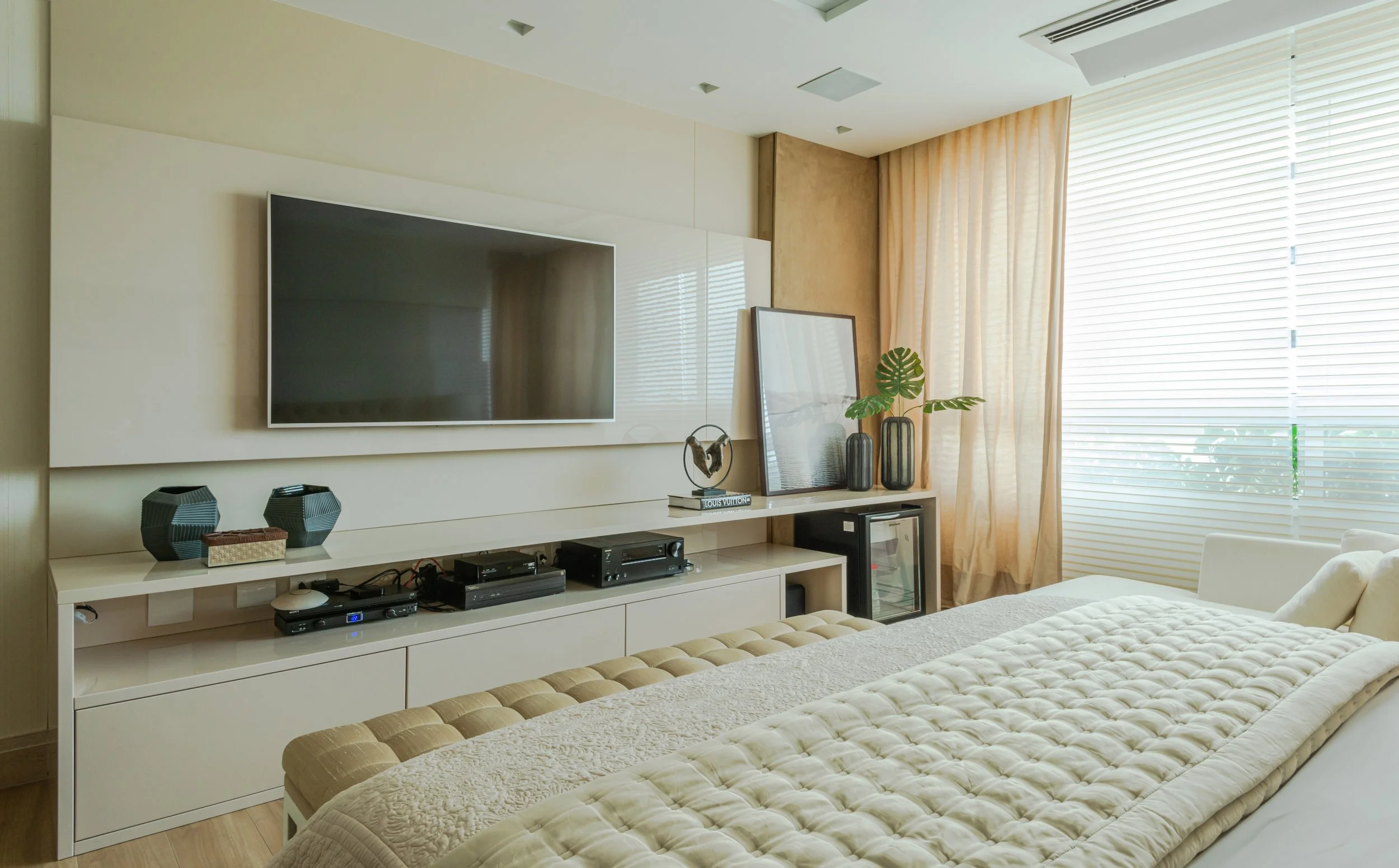 Modern hotel room with a large flat-screen TV mounted on a beige wall, a white entertainment console with electronic devices, decorative vases, framed art, and a green plant. Part of a bed with a beige quilted blanket is visible, with large windows w