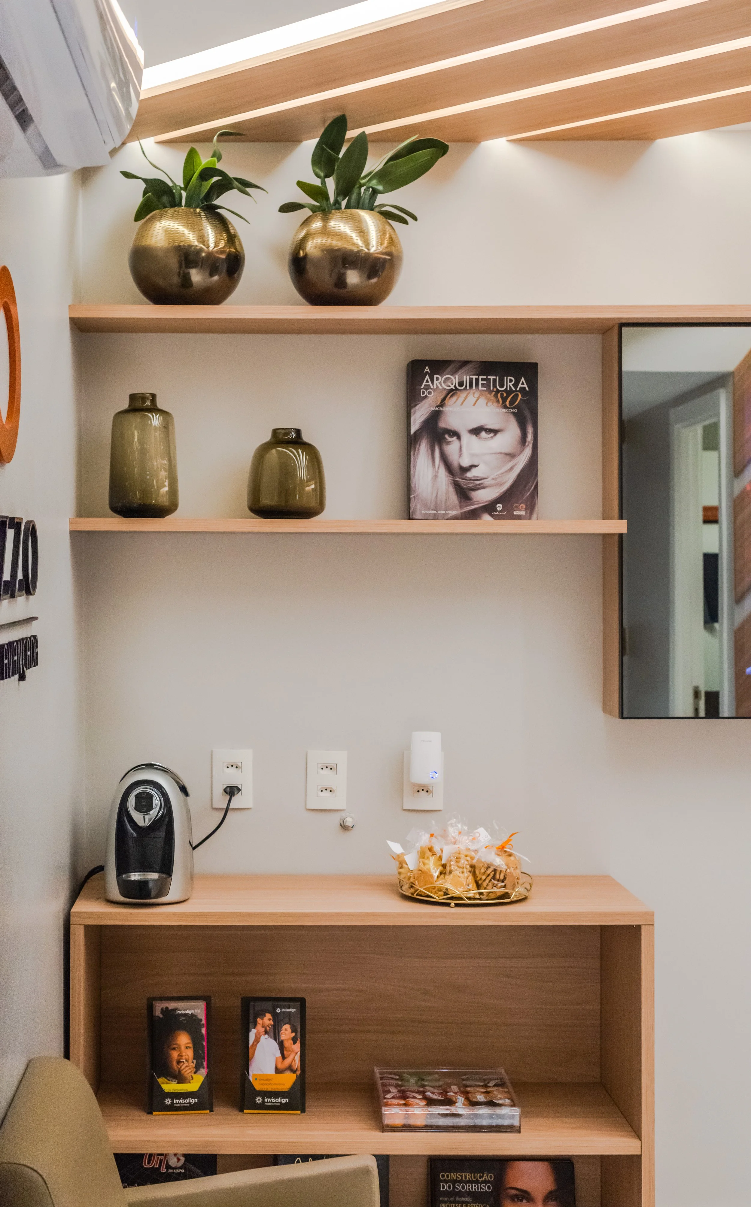Decorative shelves with green plants in metallic pots, a book titled 'A Arquitetura do Interior,' two green vases, a coffee machine, snacks on a tray, and promotional cards for Invisalign, in a room with electrical outlets and a mirror.