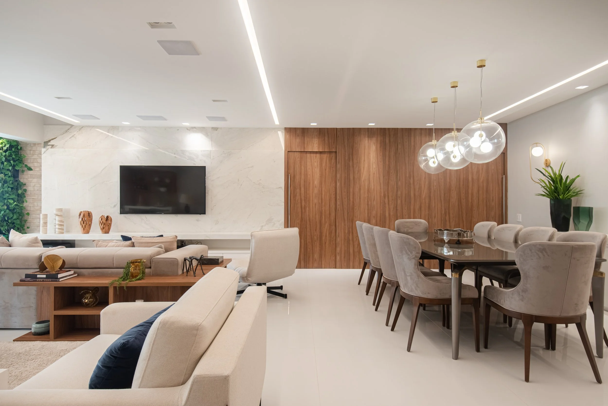 Modern living and dining room with beige sofa, armchair, flat-screen TV on marble wall, wooden dining table with chairs, glass pendant lights, indoor plant.