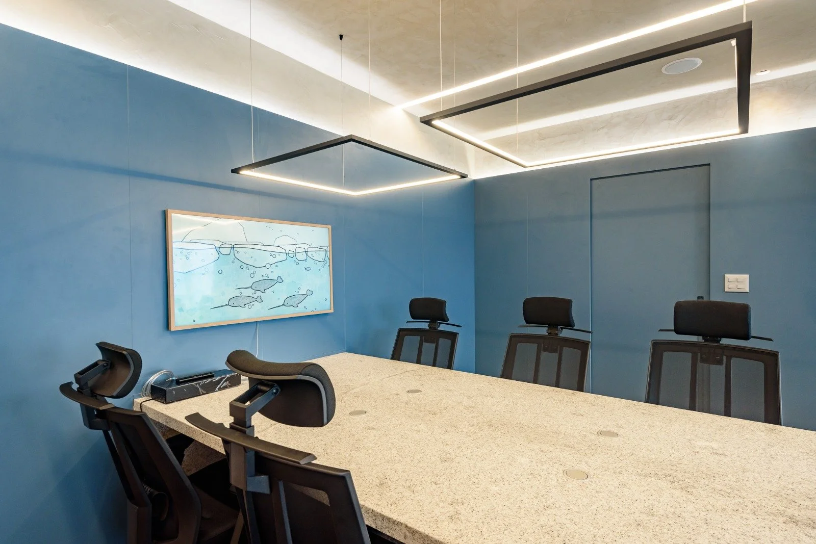 A modern conference room with a large beige marble table, black office chairs, a blue wall with a framed fishing-themed art print, and contemporary ceiling lights.