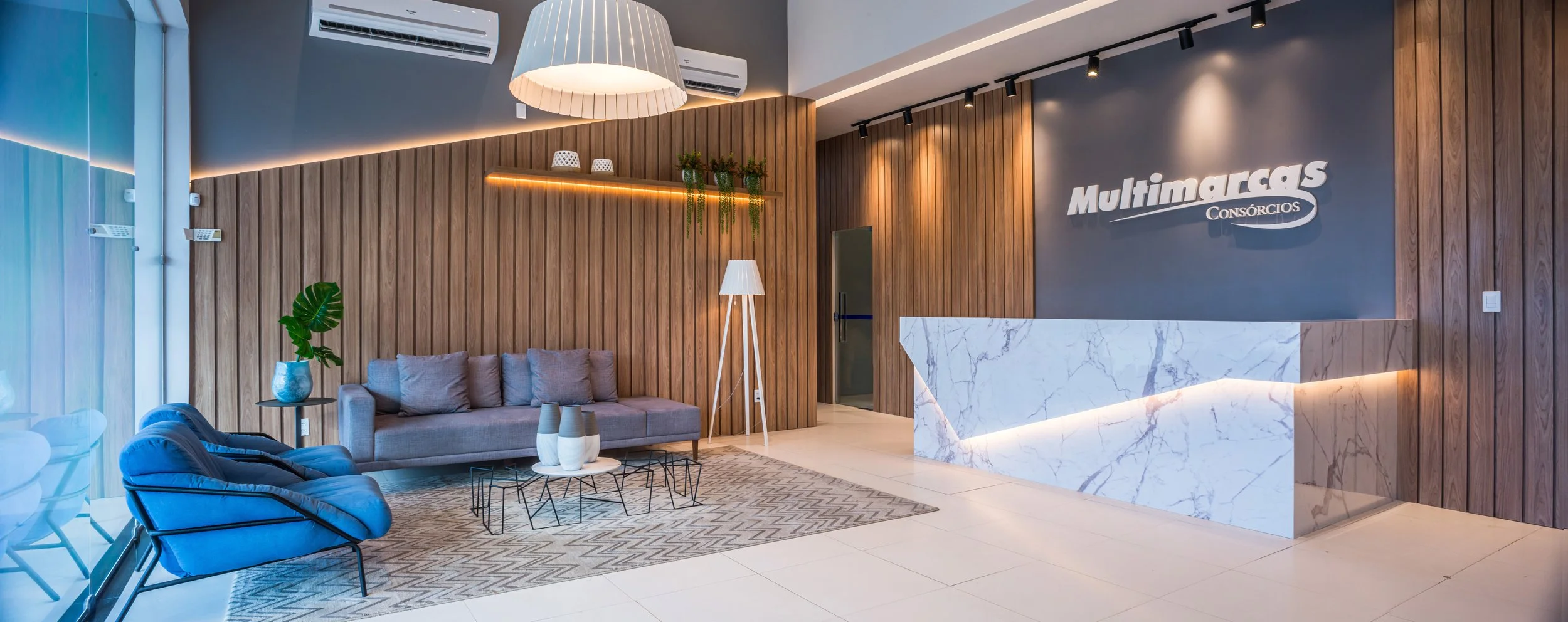 Modern office lobby with wooden walls, gray and blue seating, a white marble reception desk, and a sign reading 'Multimarcas Consórcios' on the wall. Decor includes potted plants, vases, a floor lamp, and contemporary lighting.