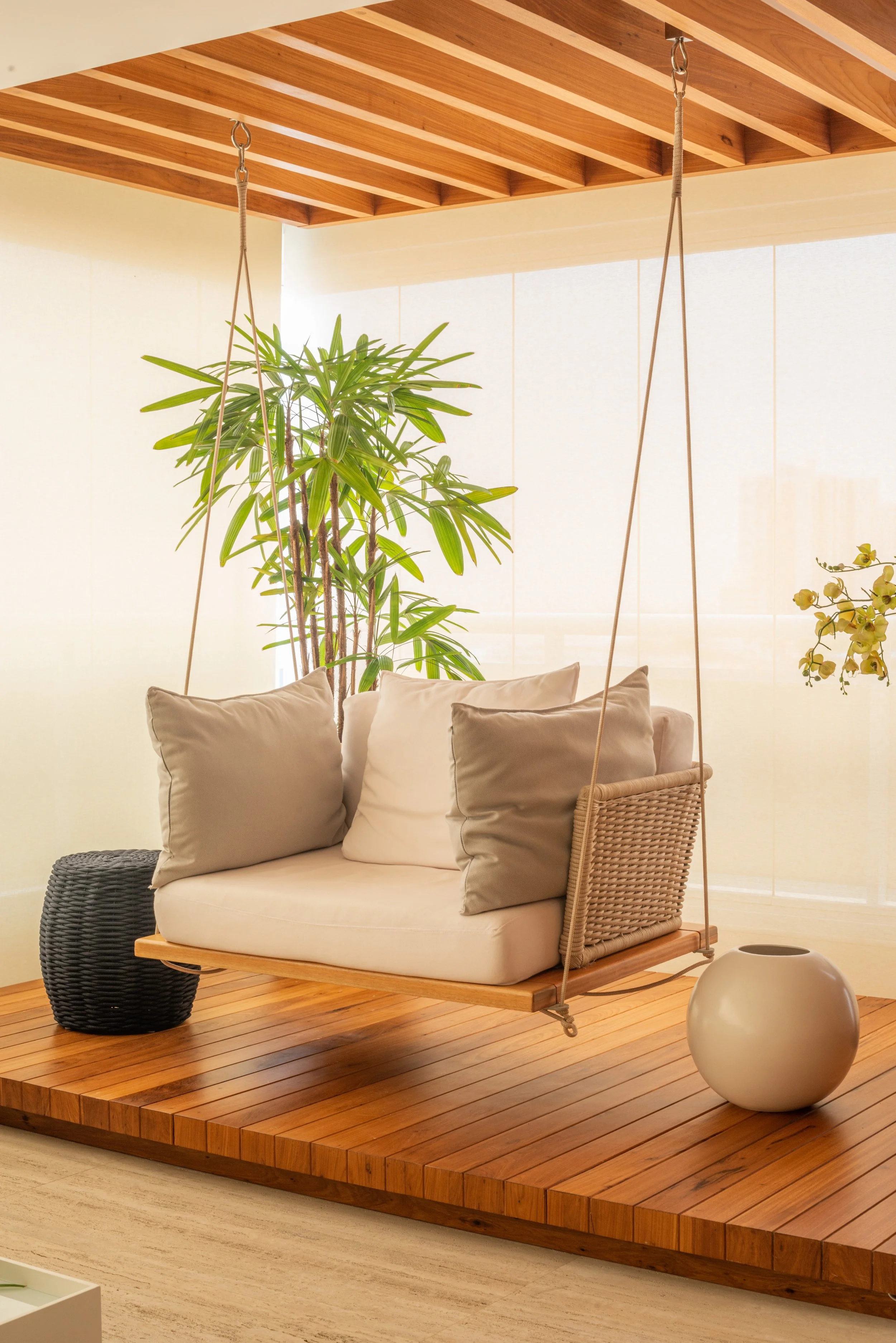 A wooden porch swing with beige cushions hanging in a bright indoor space near a large green plant and decorative vases.