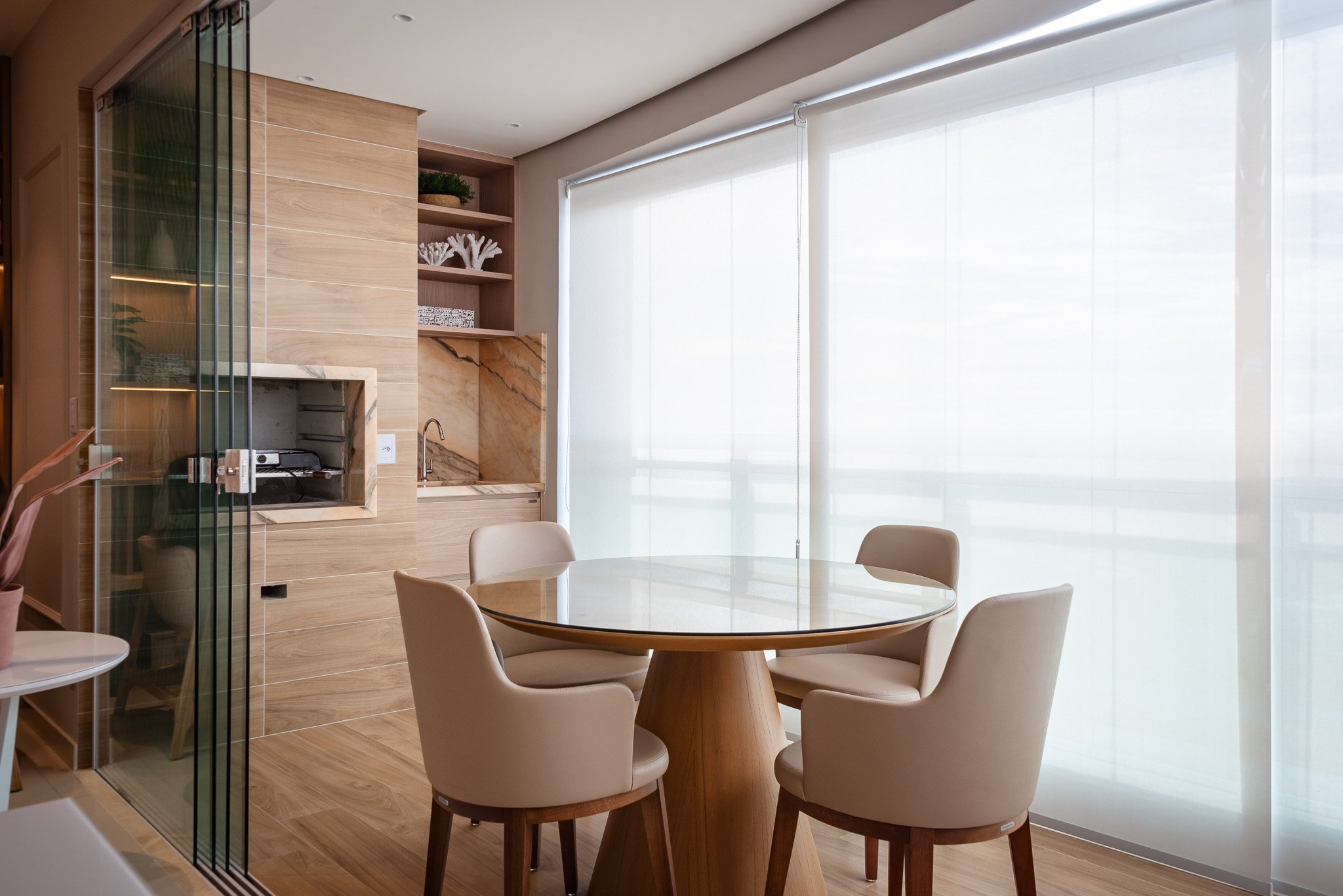Modern dining area with a round wooden table and four beige upholstered chairs, large windows with white sheer curtains, and a small kitchen nook with a sink and decorative shelves in the background.