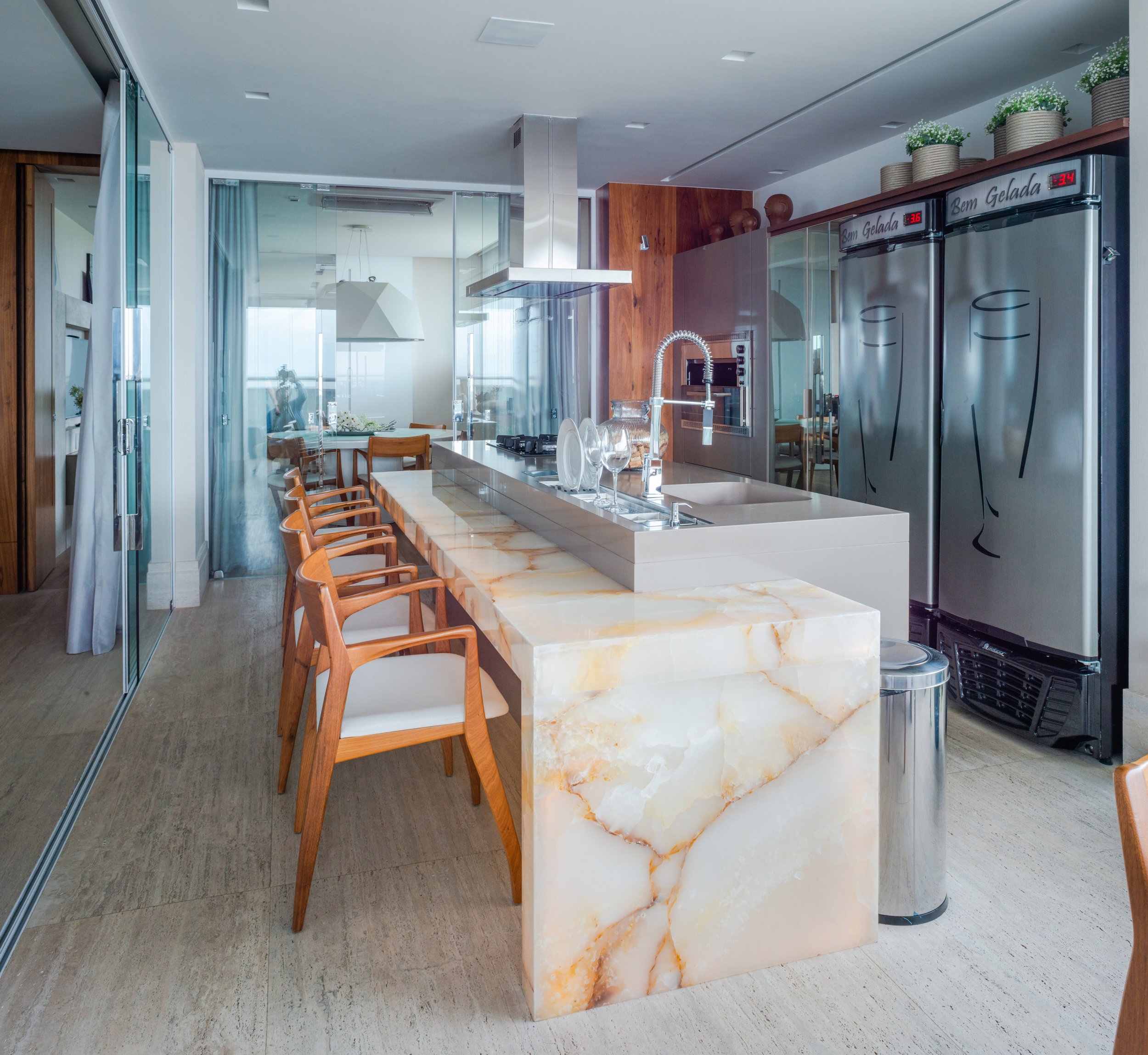 Modern kitchen with a marble island, wooden chairs, stainless steel appliances, and a glass-enclosed dining area.