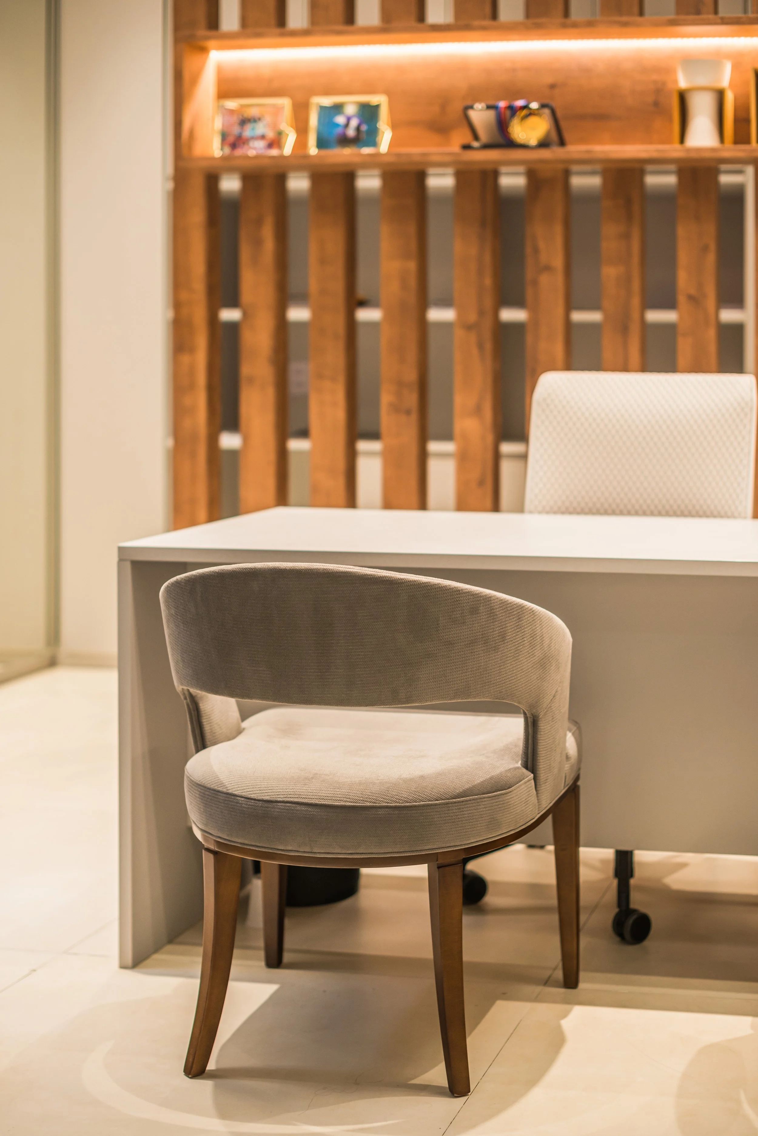 A beige upholstered chair with wooden legs in front of a white desk, with a wooden slat wall and shelves with framed photos and decorative items in the background.