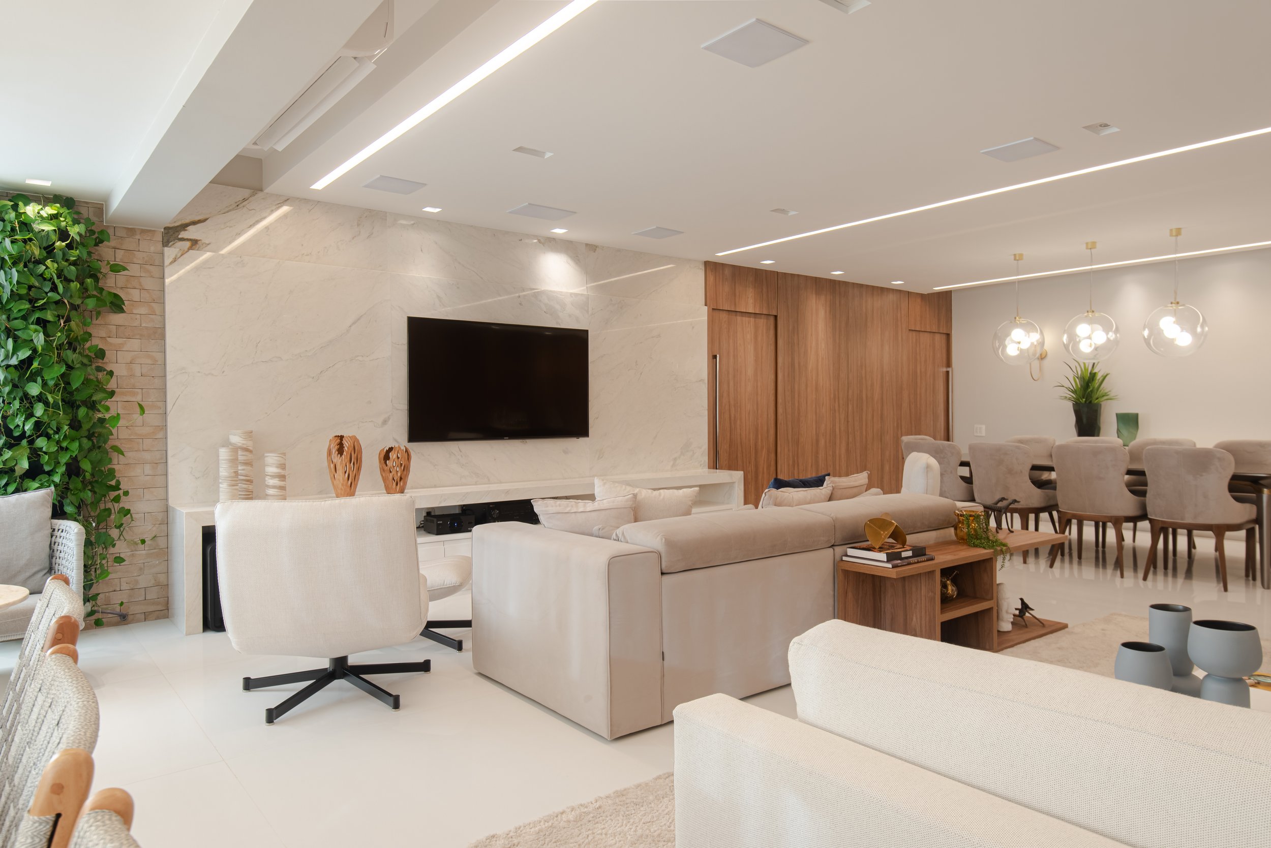 Modern living room with beige sofas, a black flat-screen TV mounted on a marble wall, green plant on the left side, a wooden hidden door, and a dining area with gray chairs and pendant lights.