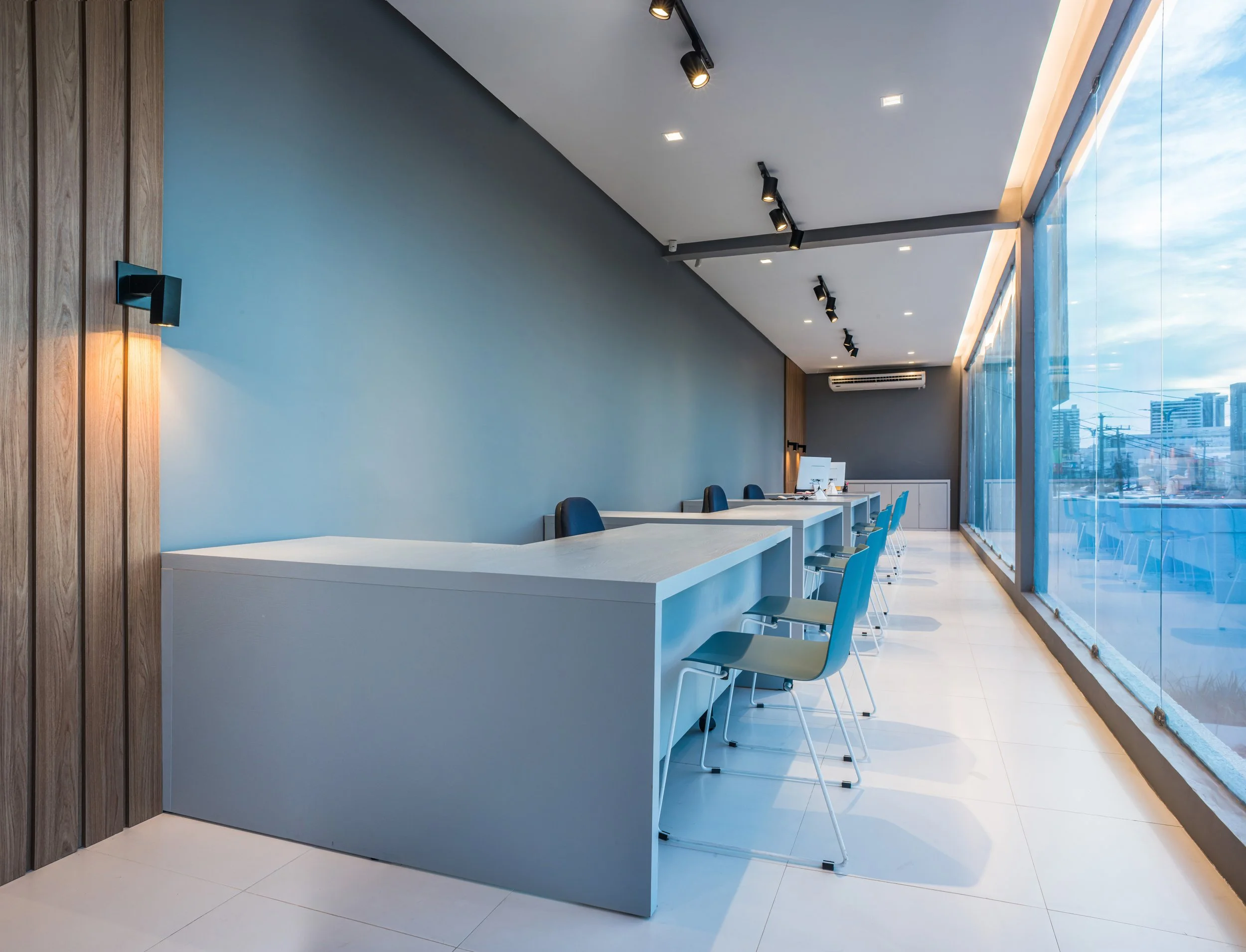 Modern office reception area with a long white counter, blue chairs, a blue wall, wood panel accents, and large floor-to-ceiling windows showing a cityscape.