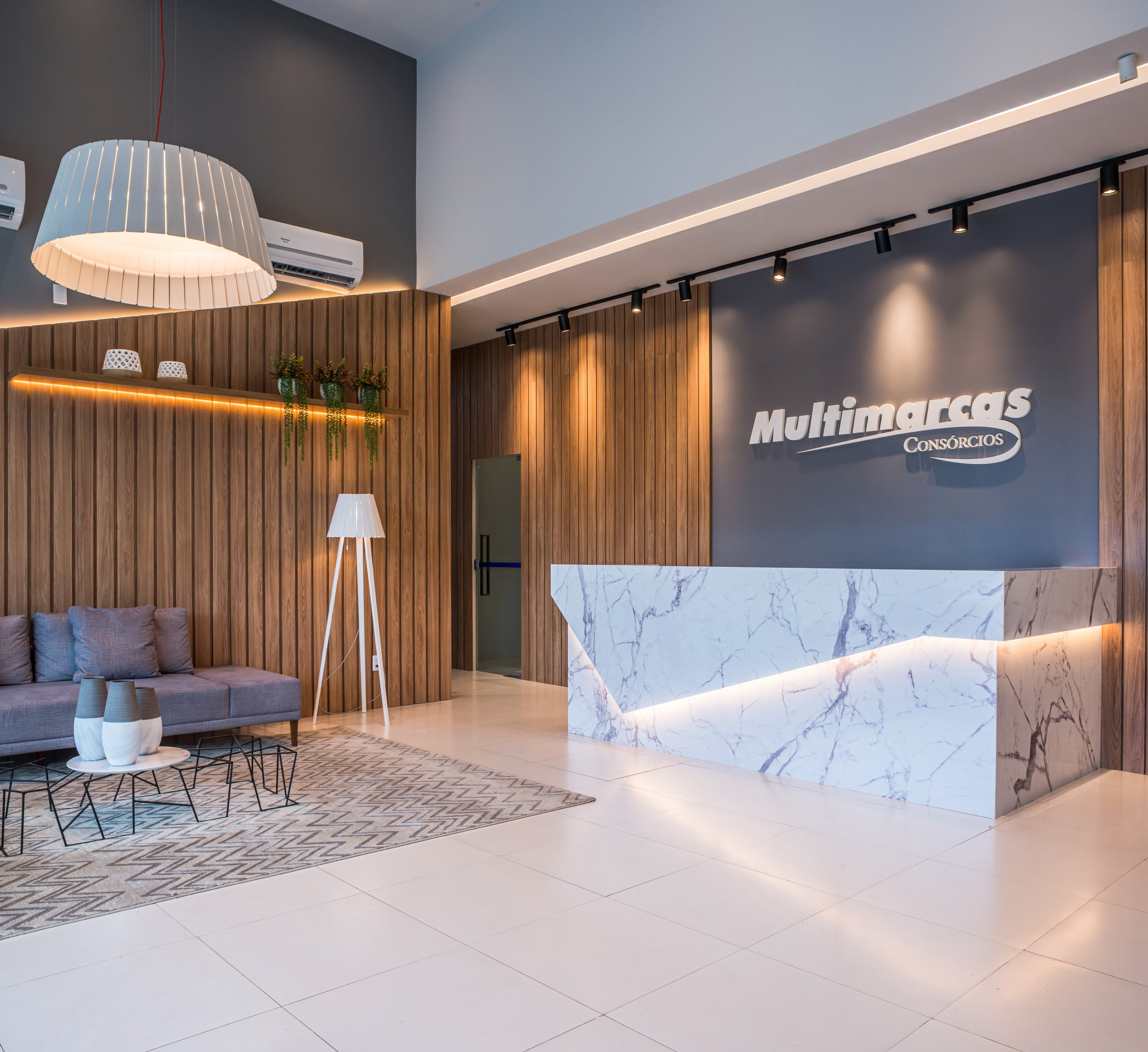Modern office reception area with wooden wall panels, a marble front desk, gray sofa, patterned rug, floor lamp, pendant light, and potted plants, with the sign 'Multimarcas Consórcios' on the wall.