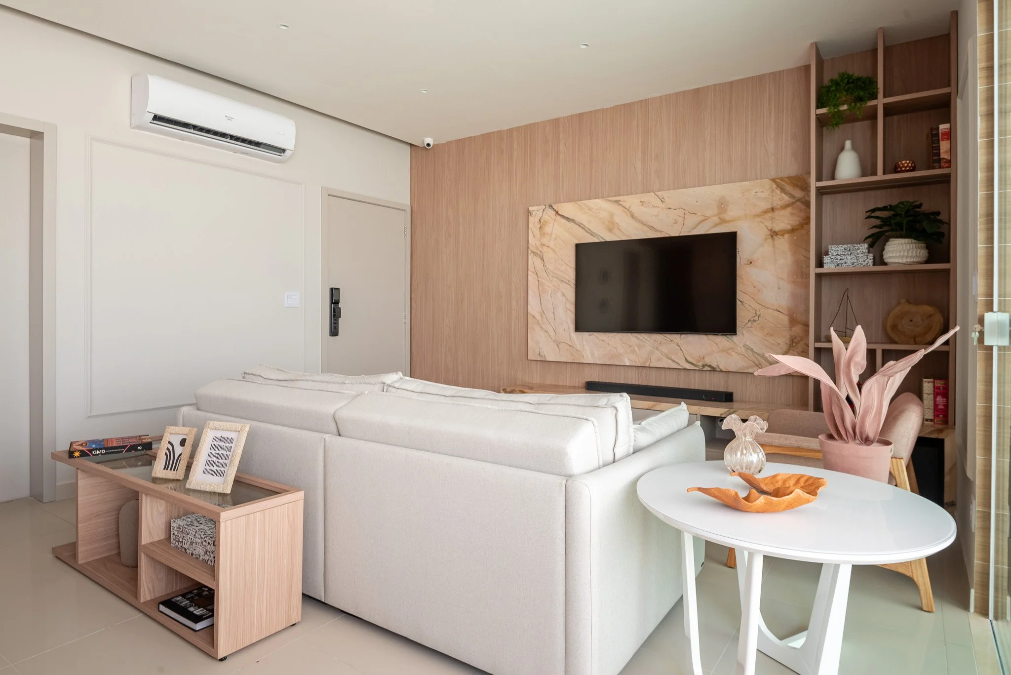 Modern living room with a white sofa, a mounted flat-screen TV on a marble accent wall, a wooden built-in bookshelf with decorative items, a white round table with a pink plant, and a wooden side table with framed photos.