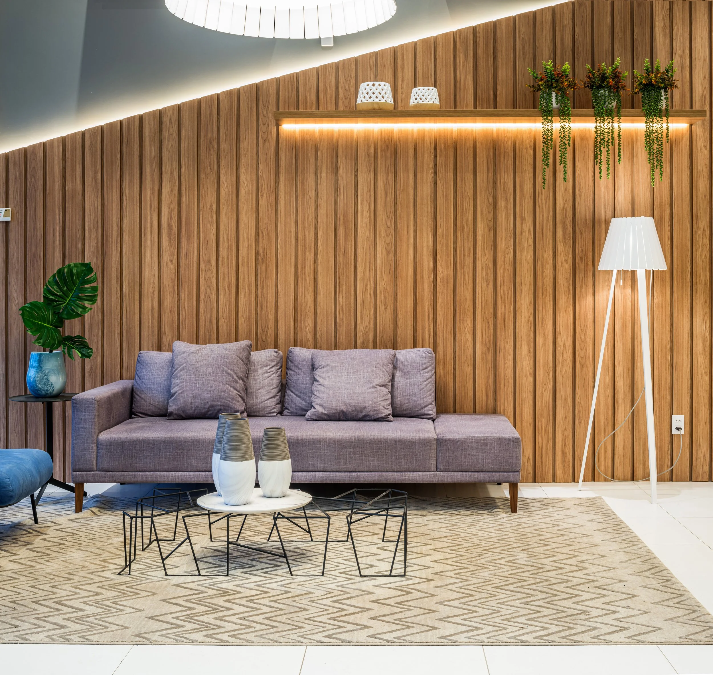 A modern living room with a purple sofa, patterned beige area rug, black metal coffee tables, potted plant on side table, wall-mounted wooden shelf with white decorative items and hanging greenery, white tripod floor lamp, and a wood-paneled wall.