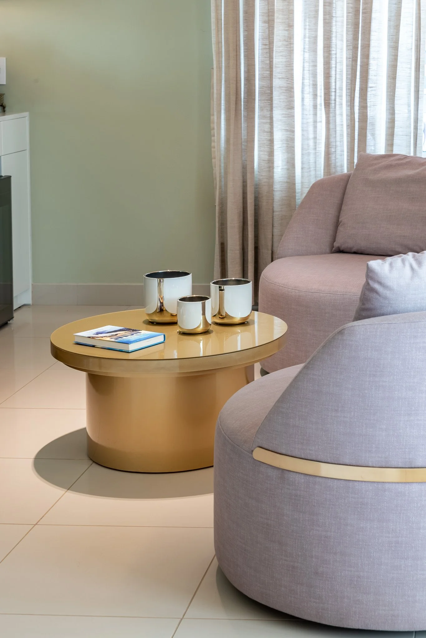Living room with beige round coffee table, books, and white and gold decorative cups, beige and pink armchairs, light green wall, beige curtains, and part of a kitchen visible