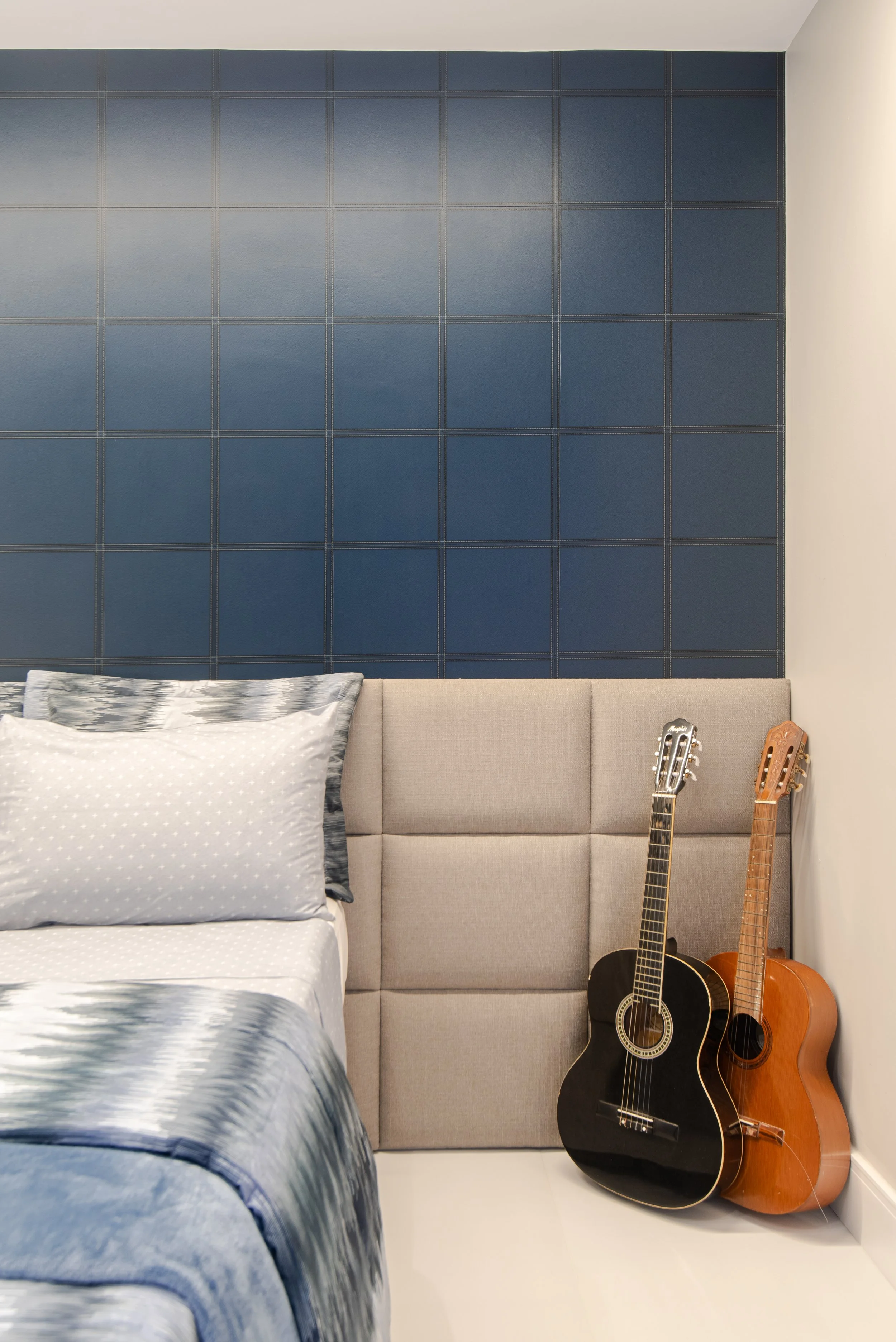 A bedroom with a beige upholstered headboard, blue wall tiles behind the bed, and two guitars leaning against the wall on the right side.