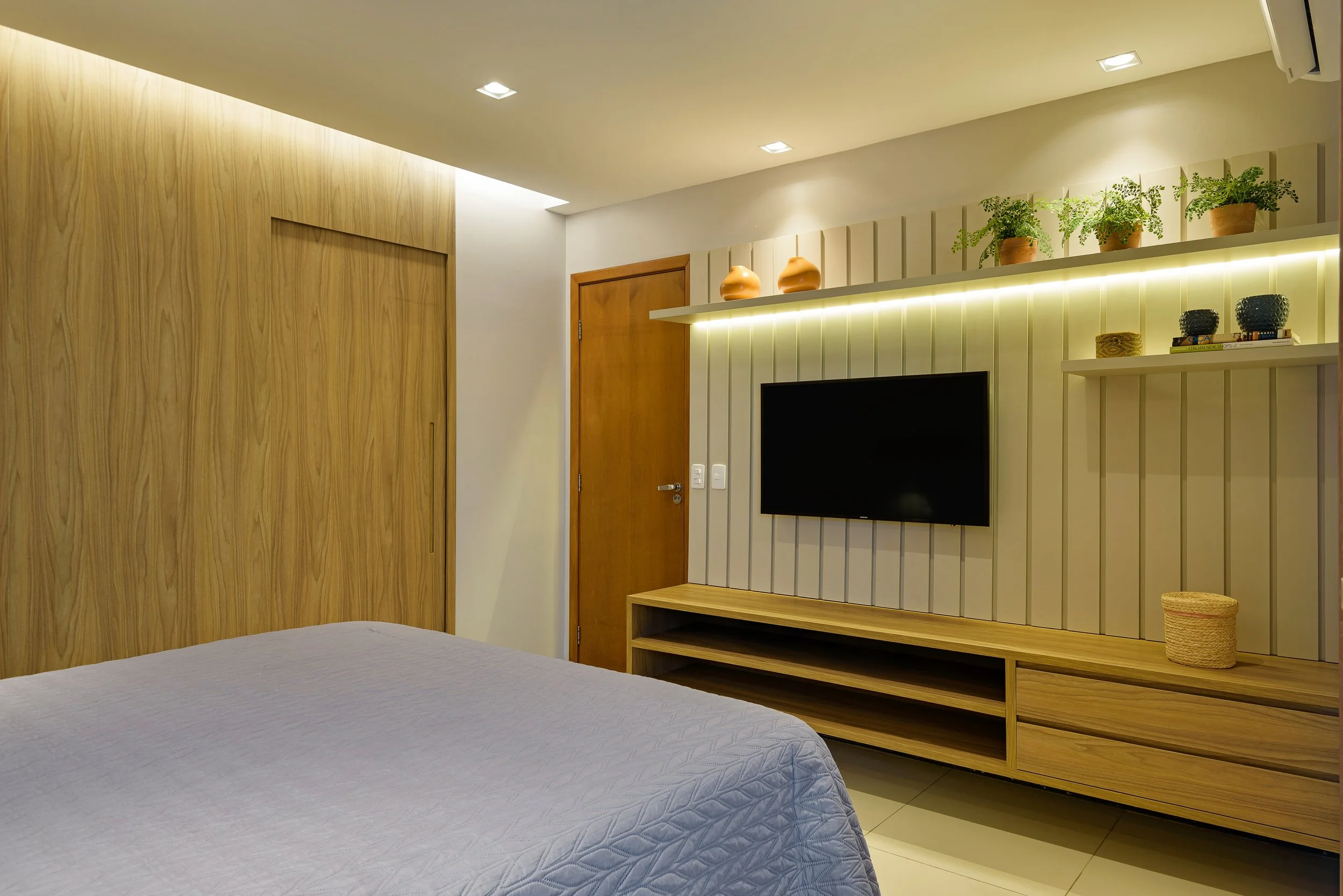 A bedroom with a bed, a wooden wardrobe, a wooden TV stand, and a wall-mounted flat-screen TV. Shelves above the TV hold potted plants and decorative objects. The room has warm wood accents and recessed ceiling lights.