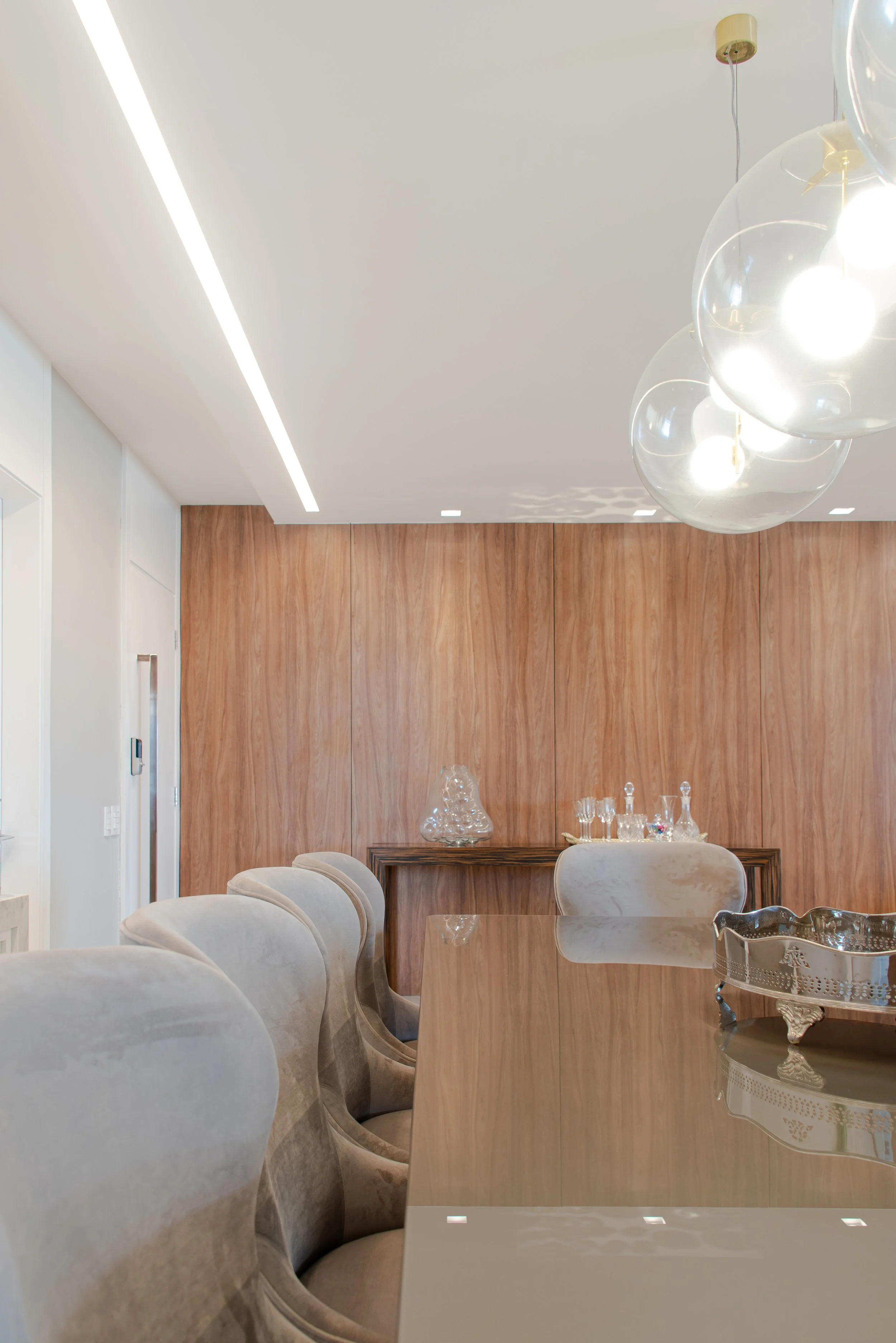 Modern dining room with beige upholstered chairs, a wooden wall, and a glossy table surface with decorative glassware and a silver tray. Bright lighting fixtures hang from the ceiling.