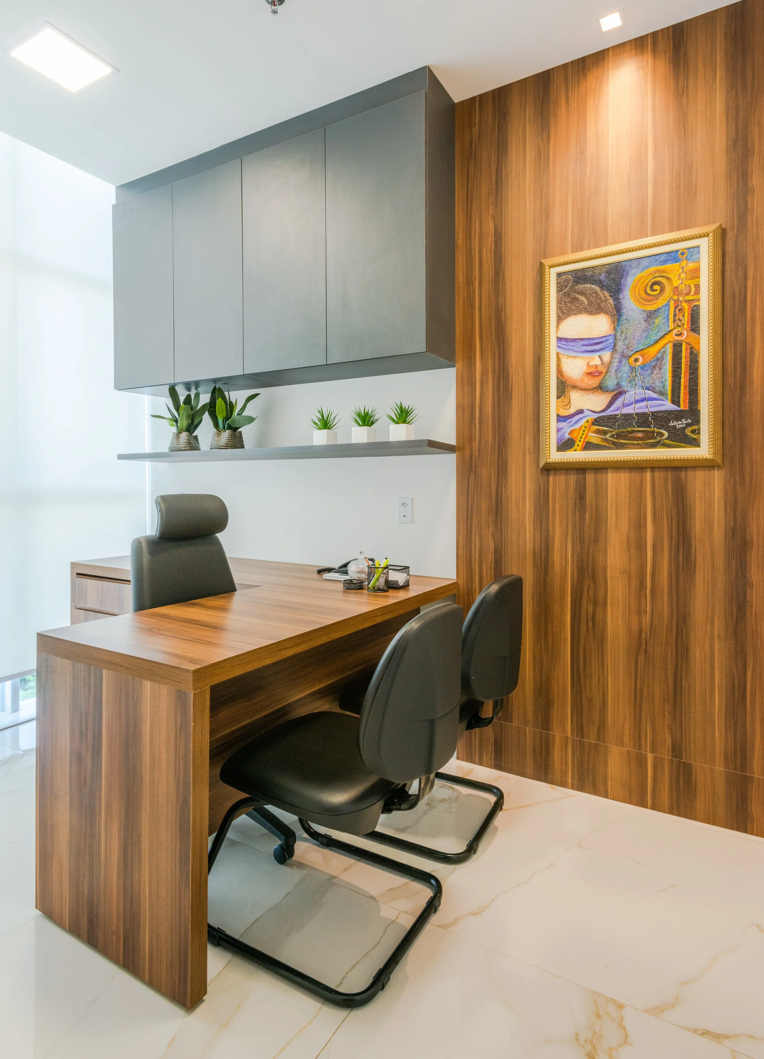 An office with a wooden desk, black chairs, wall-mounted gray cabinets, a shelf with green potted plants, and a colorful painting on a wooden paneled wall.