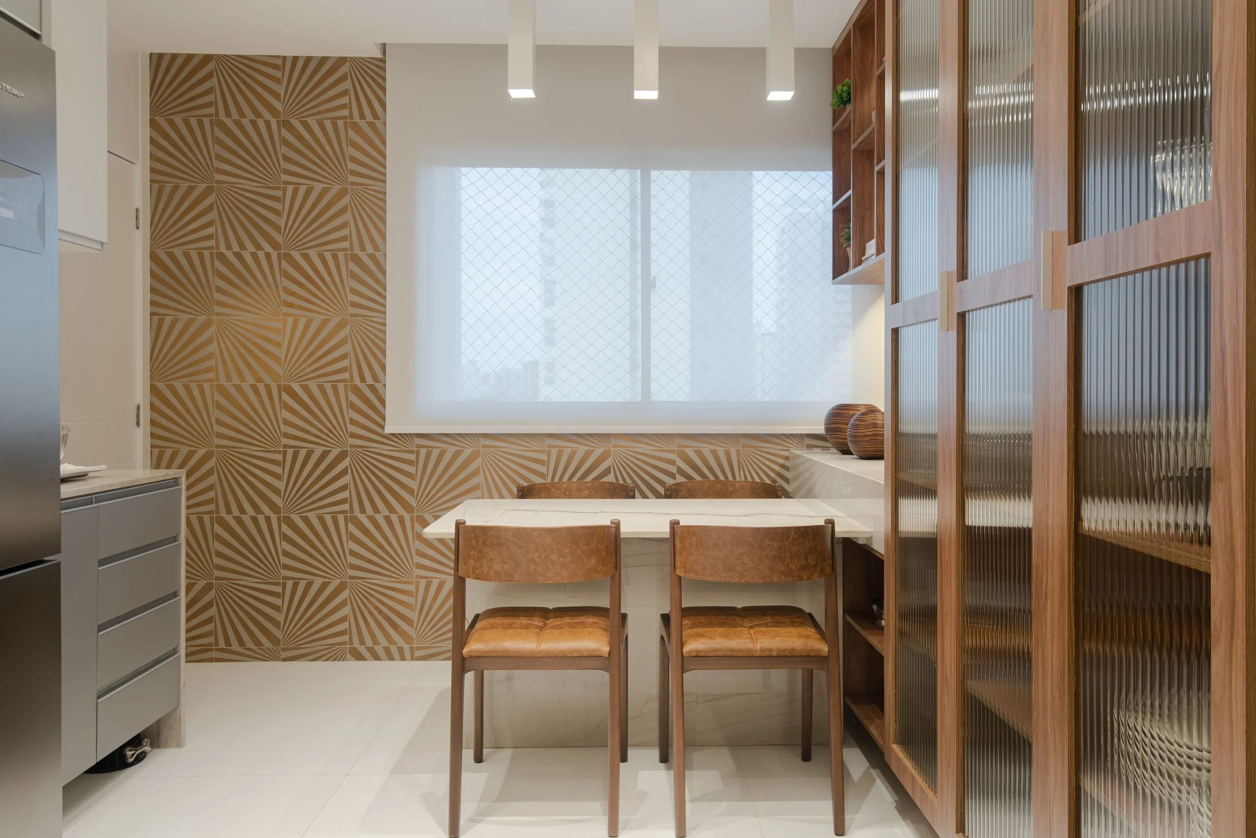 Modern kitchen with geometric patterned wall, white window with a grid and blinds, wooden cabinetry with frosted glass doors, a small dining table with three brown leather chairs, and decorative vases.